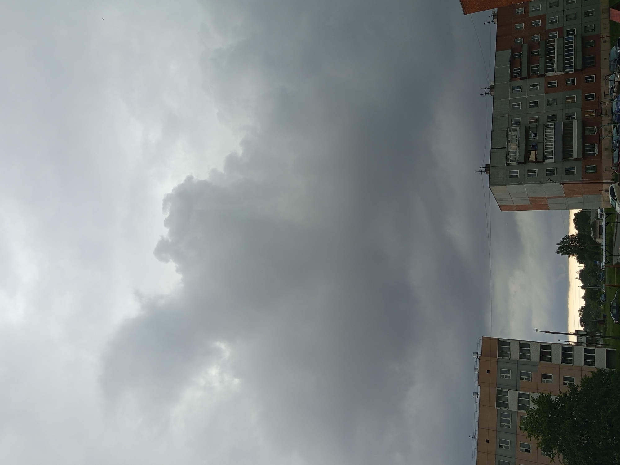 The image depicts a cloudy sky over a residential area in Gdov, Russia. Two multi-story apartment buildings are visible in the foreground, one on the left with beige and grey facades and the other on the right with grey and brick-red facades. A mature tree with dense green foliage is situated to the left of the first building, partially obscuring its lower floors. The sky is dominated by heavy, dark grey clouds, suggesting overcast weather or an approaching storm. A few scattered cars are parked on a road in the background, and a power line stretches horizontally between the two buildings. The overall impression is that of a typical urban landscape under a dramatic sky.