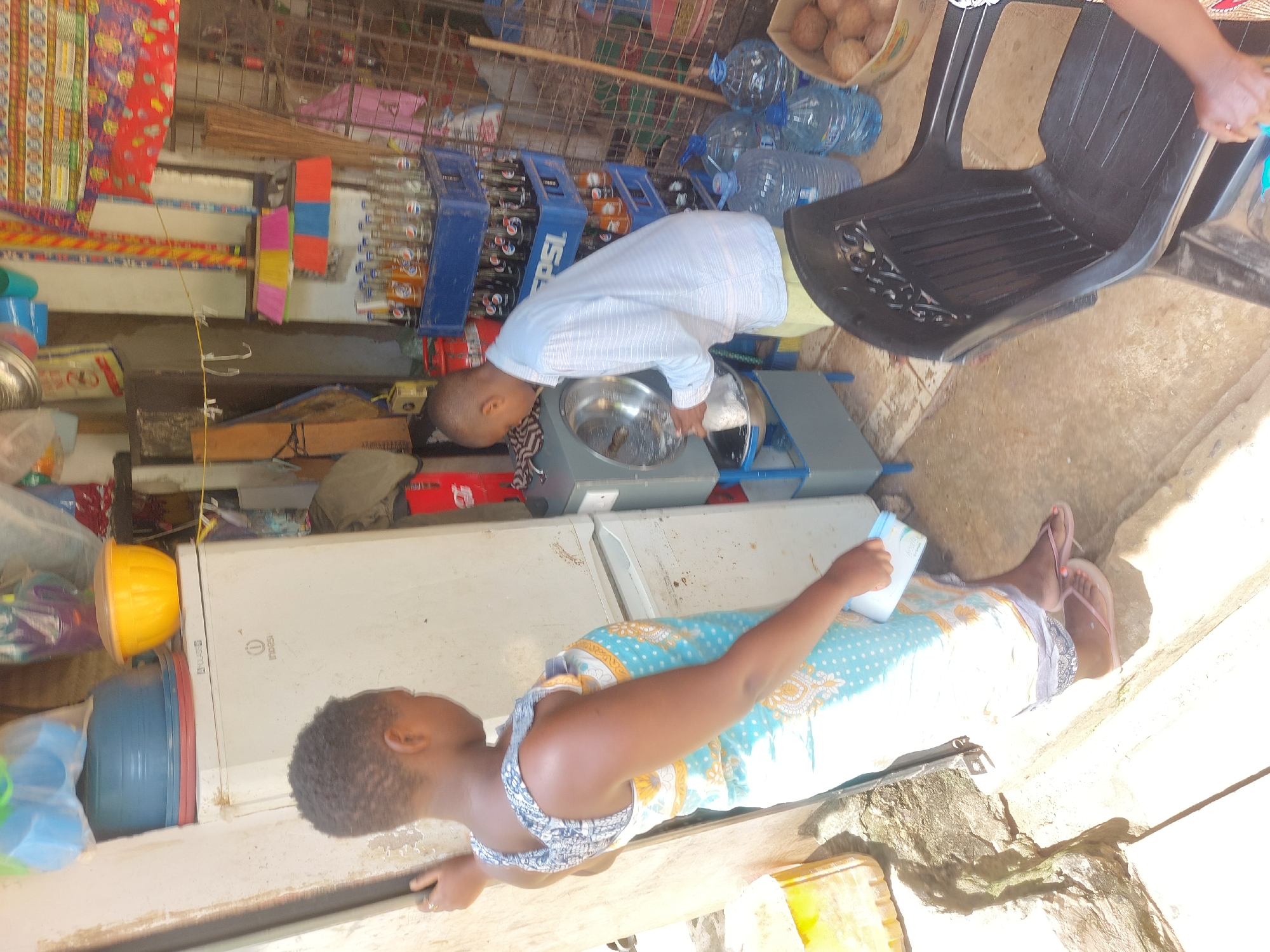 The image shows a scene within a small shop or vendor stall, likely located in a residential area. A woman wearing a blue and white patterned dress is positioned in front of what appears to be a refrigerator. She is holding a white carton. A young boy in a light blue and white striped shirt is bent over a stainless-steel bowl on a stand, appearing to be washing something, perhaps his hands or food. Behind him, crates of soft drinks (Pepsi) and bottled water are visible. Other items, including colorful plastic containers and cooking supplies, are scattered around the stall. A black plastic chair is visible, as well as a crate with what appear to be coconuts. The environment suggests a modest retail operation in a neighborhood setting. Without additional information or data points, a determination of the location in the city of Dar es Salaam is not possible.