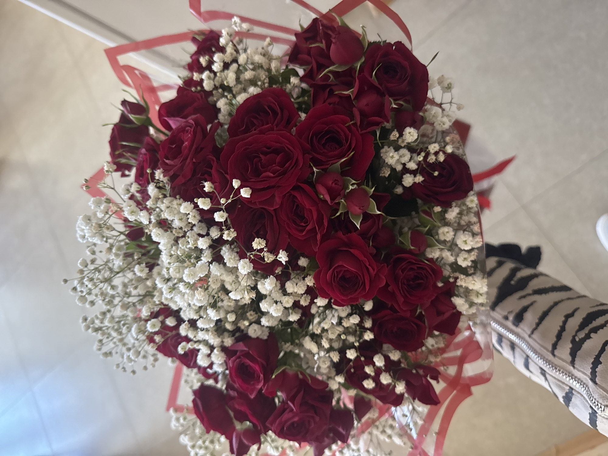 The image shows a bouquet of deep red roses interspersed with white baby's breath flowers. The bouquet is wrapped with sheer red ribbon. It is unclear what the exact location is. The presence of what appears to be a jacket or a bag with a tiger-stripe pattern in the periphery suggests an indoor setting.