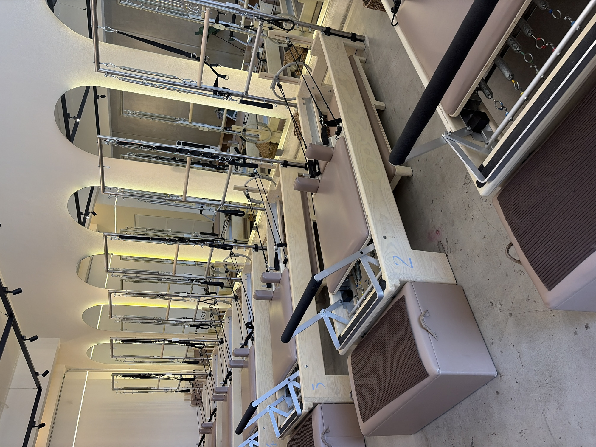 This image displays a modern Pilates studio, featuring multiple reformers lined up in rows. The scene is captured from a slightly elevated, angled perspective, looking down the length of the studio.
The primary subjects are numerous Pilates reformers, constructed with light-colored wooden frames and beige or light brown padded carriages. Each reformer is equipped with springs of various colors (red, blue, yellow, green), black straps, and padded handles or foot bars. Some reformers also have small, matching padded boxes placed on the concrete floor beside them. The reformers are arranged in an orderly fashion, suggesting a class setup.
The studio's interior design is a notable detail, characterized by a clean and contemporary aesthetic. The white ceiling features several large, arched cutouts, which are mirrored to create reflective surfaces. These arched mirrors reflect parts of the reformers and strip lights embedded within the ceiling structure, adding depth and brightness to the space. Additional black track spotlights are visible on the left side of the ceiling. The floor is made of light grey concrete, complementing the neutral color palette of the equipment and walls.
There are no people present in the image, indicating the studio is either empty before or after a class, or during off-hours. The activity implied is Pilates exercise. The studio appears well-maintained and ready for use. Faint blue numbers, specifically "2" and "5", are visible on the wooden frames of two of the reformers, likely for identification. The overall impression is one of order, cleanliness, and modern design.