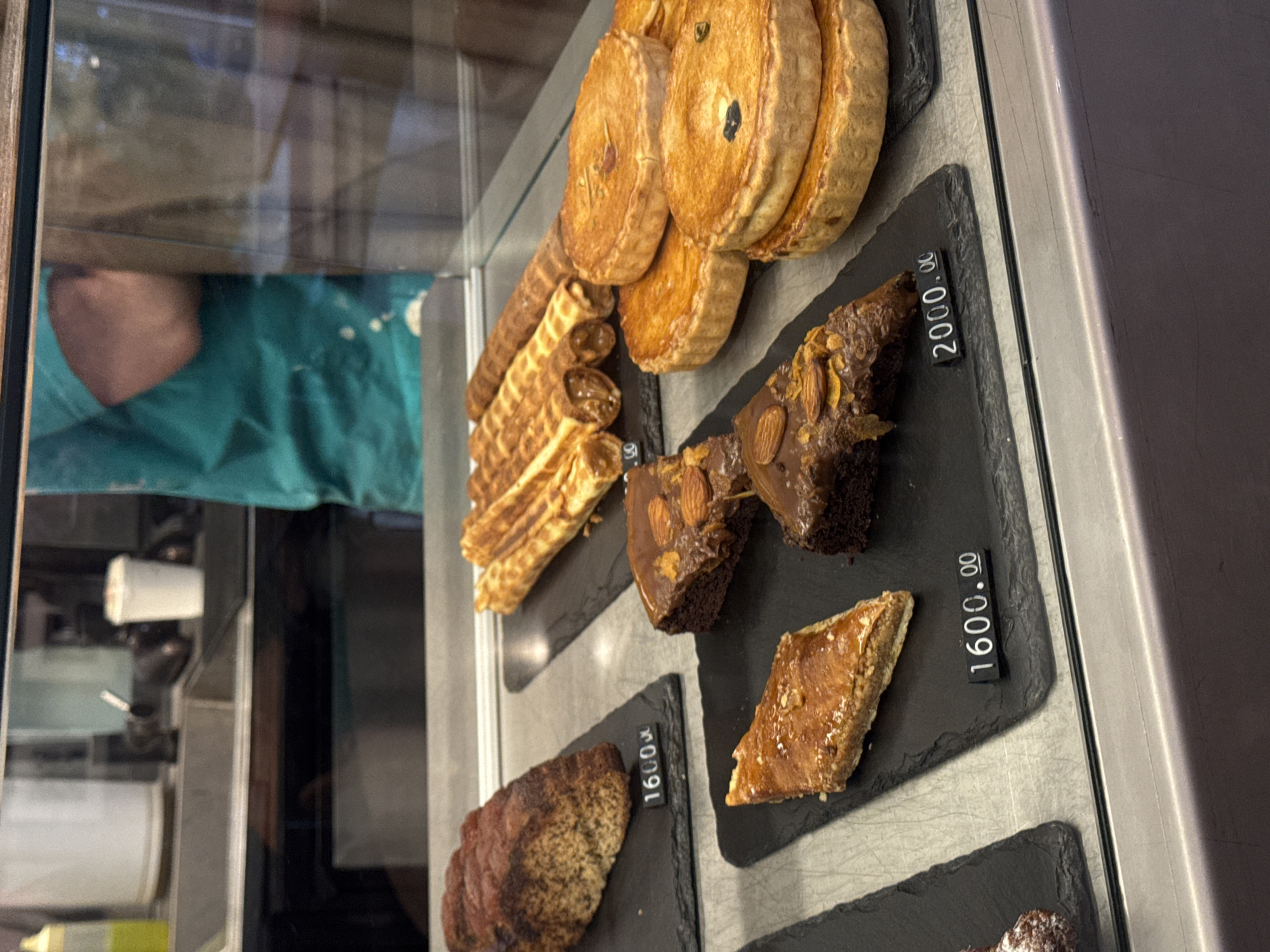 This close-up image shows a variety of baked goods arranged in a display case, likely in a bakery or cafe in Yerevan, Armenia. The pastries are presented on black rectangular serving boards.
In the upper section of the display, there are several round, golden-brown pies or tarts, some with visible dark specks suggesting a filling. Below these, a stack of golden, tubular waffle rolls, some appearing to be filled, are visible. To the right, two pieces of dark chocolate brownie are featured, generously topped with sliced almonds and a glossy glaze. A small black price tag next to these brownies clearly shows "2000.⁰⁰" with the Armenian Dram () currency symbol.
Further down, on another black serving board, a piece of golden, flaky pastry, possibly baklava or a similar honey cake, with visible nuts, is displayed. Adjacent to it, a price tag reads "1600.⁰⁰" also accompanied by the Dram currency symbol. Partially visible at the bottom left is a slice of a loaf-shaped pastry, possibly a cake or banana bread, with a similar price tag also showing "1600.⁰⁰" and the Dram symbol.
Through the glass of the display case, reflections are prominent. In the upper left, a person wearing a teal or green uniform or apron is partially reflected. The setting appears to be an indoor commercial space, with professional presentation of the food items for sale.