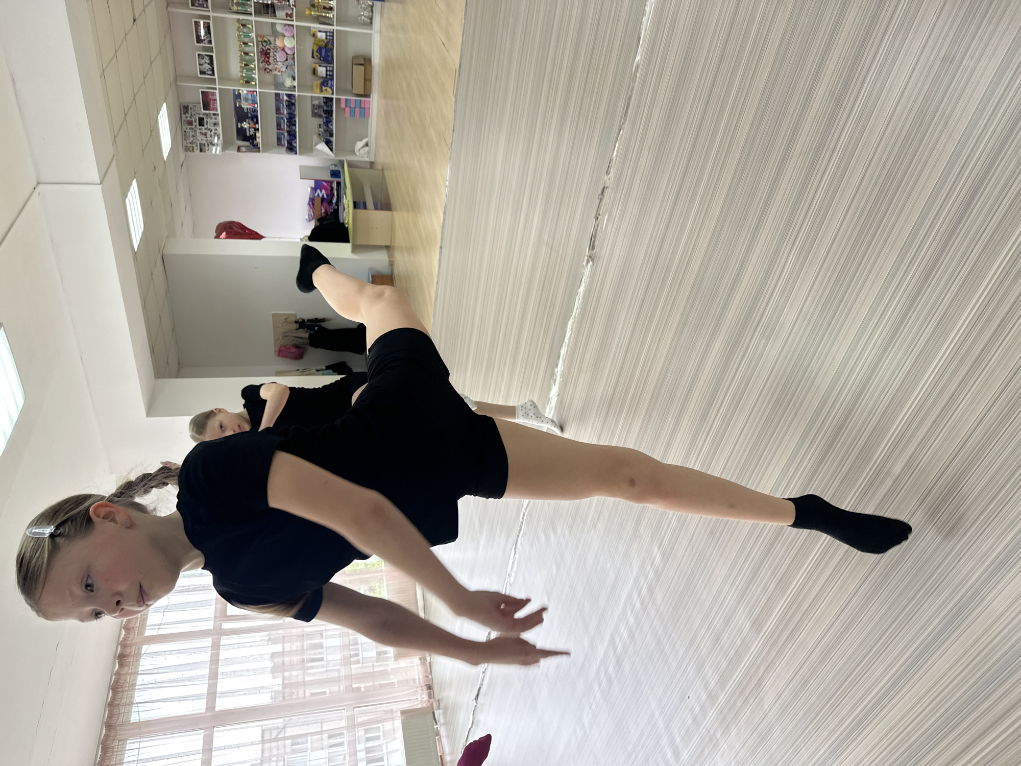The image shows a young girl in a black shirt and shorts performing a stretching exercise in what appears to be a dance or fitness studio. Her left leg is extended forward on the floor while her right leg is lifted upwards. She is wearing black socks. Another person is partially visible behind her, also dressed in black. The floor is light-colored with a striped pattern. In the background, there are shelves with various items, possibly trophies or decorations. The presence of large windows in the scene suggests that the location is likely indoors. The location in the city of Votkinsk, Russia, cannot be determined with the current image.