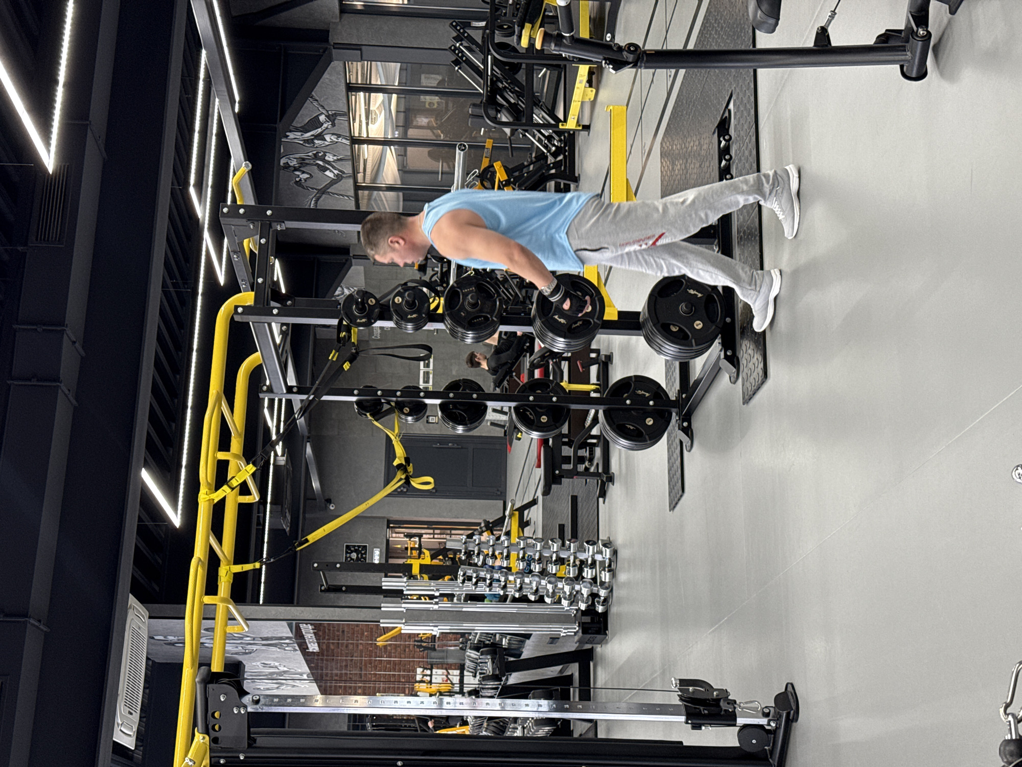 The image captures an indoor scene within a gym facility, possibly in Votkinsk, Russia, though no location-specific elements confirm this directly. A man wearing a light blue t-shirt and light gray sweatpants is performing a weightlifting exercise. He is positioned near a weight rack, holding a barbell loaded with weight plates. The rack itself is black and yellow and filled with multiple sets of weights. Other gym equipment, including dumbbells and cable machines, can be seen in the background, indicating a fitness-oriented environment. The gym's interior design features a combination of black and gray elements, accented with bright yellow equipment components and linear LED lighting fixtures. The floor is a light gray concrete, and a large mirror covers a portion of the wall.