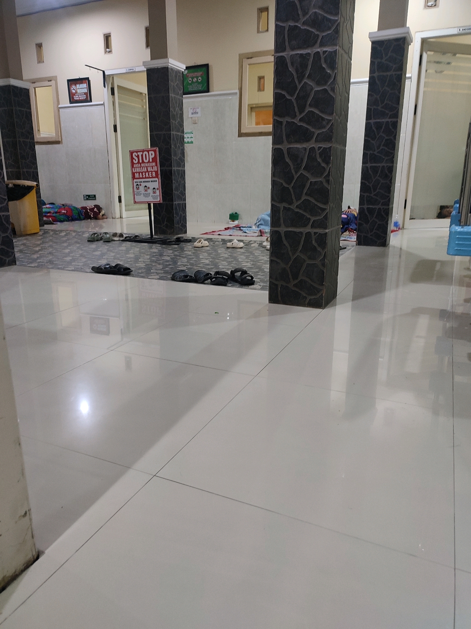 The image depicts an interior hallway with a tiled floor and patterned pillar structures. A person is lying down on the floor in the background, covered by a blanket. There are several pairs of sandals scattered on the floor. A red and white sign that reads "STOP ANDA MEMASUKI KAWASAN WAJIB MASKER" (STOP YOU ARE ENTERING A MASK REQUIRED AREA) is prominently displayed near the pillars. To the left of this sign, a framed picture with a red prohibition symbol and text "DILARANG MEROKOK" (NO SMOKING) is visible. Further to the left, a yellow trash bin is positioned near a window. Another framed sign with green symbols and text is mounted on the wall in the background. The environment appears to be an indoor public space, possibly a medical facility or a waiting area, located in Sukorejo, Indonesia, given the Indonesian text on the signs. The lighting suggests an indoor setting, possibly during the day or under artificial light.