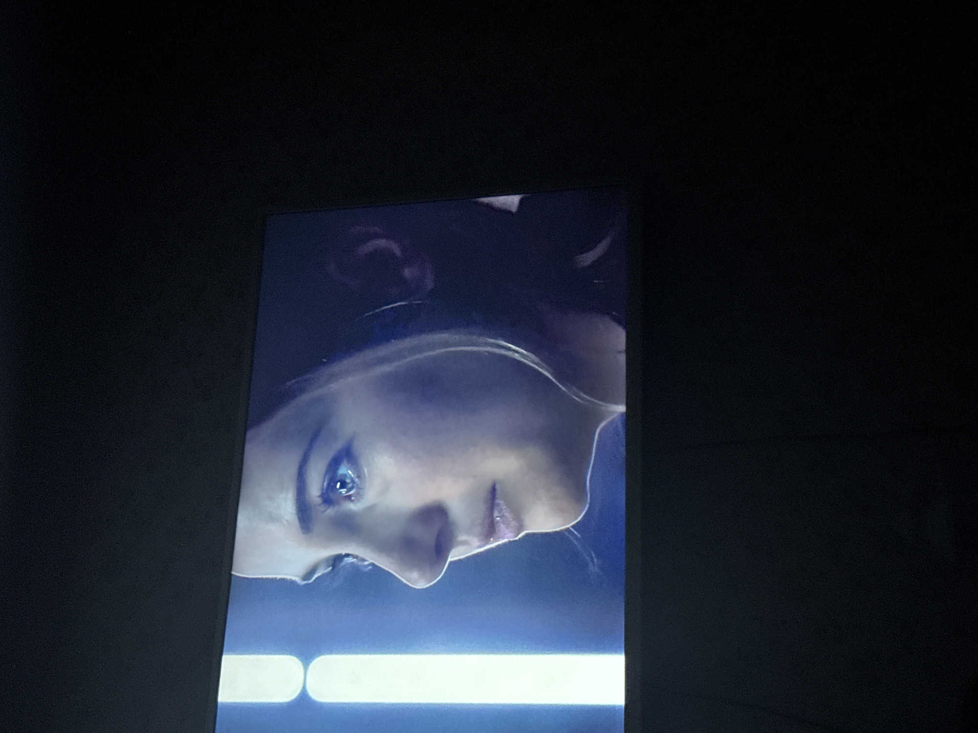 The image shows a close-up of a woman's face, illuminated by a blue light. The image appears to be displayed on a screen or monitor. The surrounding environment is dark. There is no indication of Mytishchi, Russia, or any other specific location.