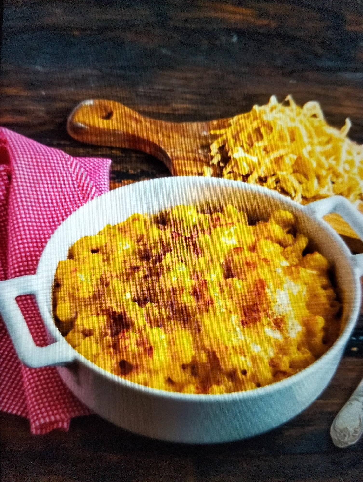 A white oval bowl filled with macaroni and cheese sits on a dark wooden surface. A red and white checkered napkin is placed to the left of the bowl. To the right of the bowl, a wooden spoon holds a pile of shredded cheese. A fork with an ornate handle rests on the right side of the wooden surface. The scene is lit from above, casting subtle shadows. There are no people present in the image. The location in the city of Payagaboh, Indonesia is not determinable from the visual information provided.