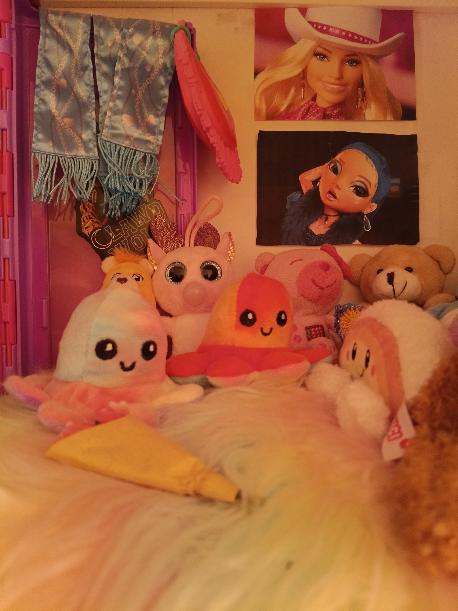 The image shows a collection of stuffed animals arranged on a soft, fluffy surface. In the background, there are two framed pictures of dolls and a light blue scarf with a chain pattern hanging on a pink structure. A pink accessory is also visible on the right side of the scarf. One stuffed animal is a white octopus with black eyes and a smiling mouth. Another is a pink and white stuffed animal with large, sparkly eyes and pink ears. A small, yellow bear is peeking out from behind it. There is also a pink stuffed dog and a brown teddy bear. Another stuffed animal is orange and has a smiling face. A yellow, cone-shaped object lies in the foreground. The overall setting appears to be indoors, possibly a child's room or a display. There is no indication of the city of Wels, Austria.