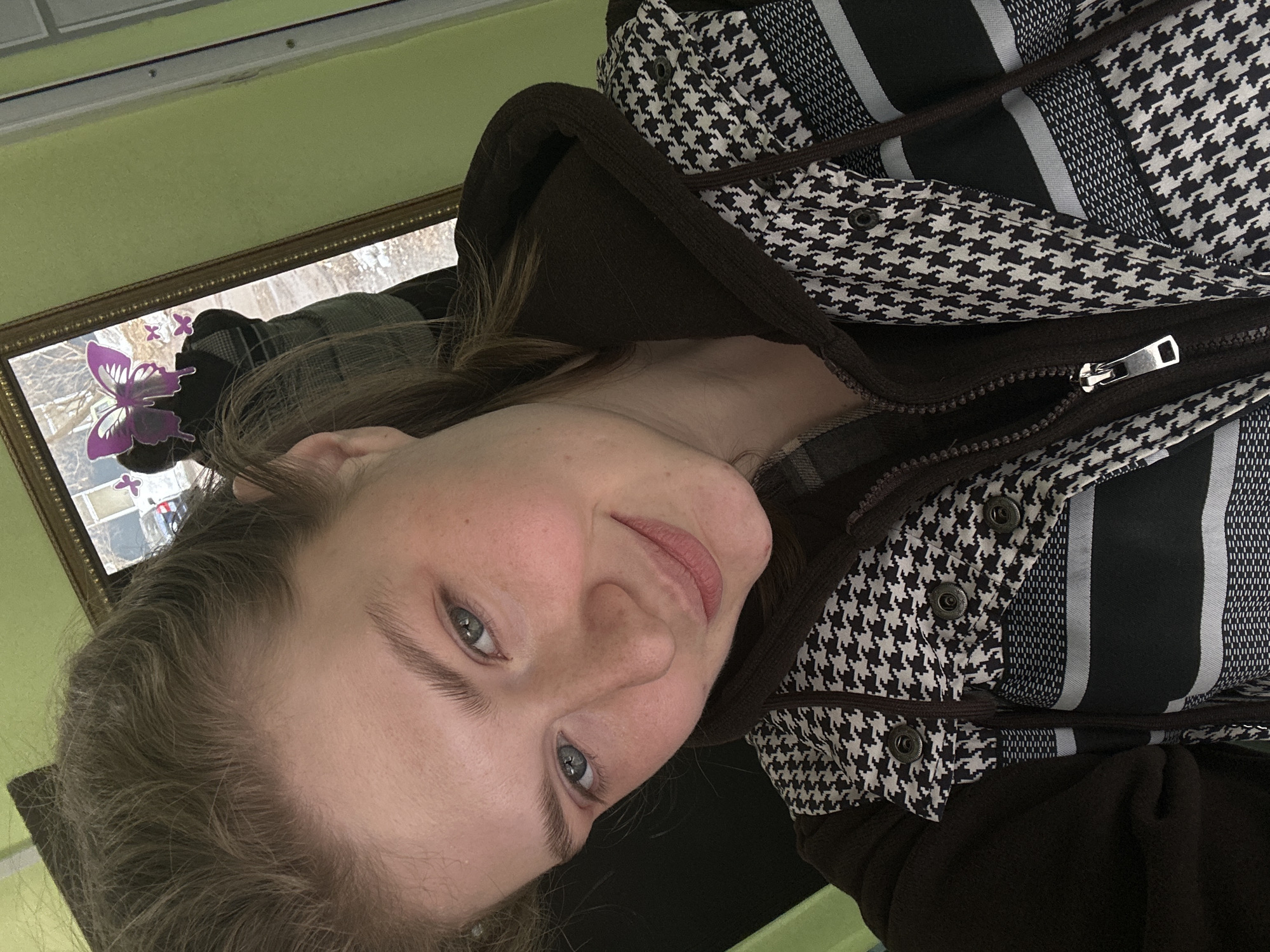 The image displays a close-up indoor selfie of a young adult female, positioned against a light green wall. The subject has light brown hair, blue-grey eyes, fair skin with visible freckles, and a subtle upward turn to her lips. She is wearing a dark brown hooded garment beneath an outer layer that features a black and white houndstooth pattern, horizontal black and grey stripes, and metallic snap fasteners. A partially unzipped silver zipper is also visible on the outer garment. Behind her right shoulder, a gold-framed mirror reflects an outdoor scene, indicating a cold season with patches of snow on the ground, a light-colored building, and a dark vehicle. Several purple butterfly-shaped decorative items are affixed to the mirror's surface, partially obscuring the reflection. A partial reflection of an individual wearing a patterned vest, consistent with the subject's attire, is also visible in the mirror. To the right of the mirror, a white vertical frame with exposed fasteners is present. The scene is located in Izhevsk, Russia.