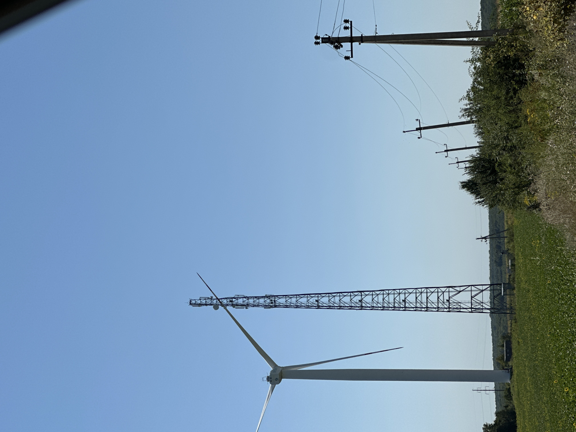 This is an outdoor, eye-level, vertical medium shot photograph taken on a sunny day in the city of Korchivka, Ukraine. The image features a field of green crops, with a single wind turbine on the left side and a tall, lattice-style telecommunications tower in the center. In the background, a line of utility poles with power lines stretches across the horizon, extending towards the right side of the frame. The sky is clear and blue. The overall scene depicts a rural landscape with infrastructure for power generation and communication.