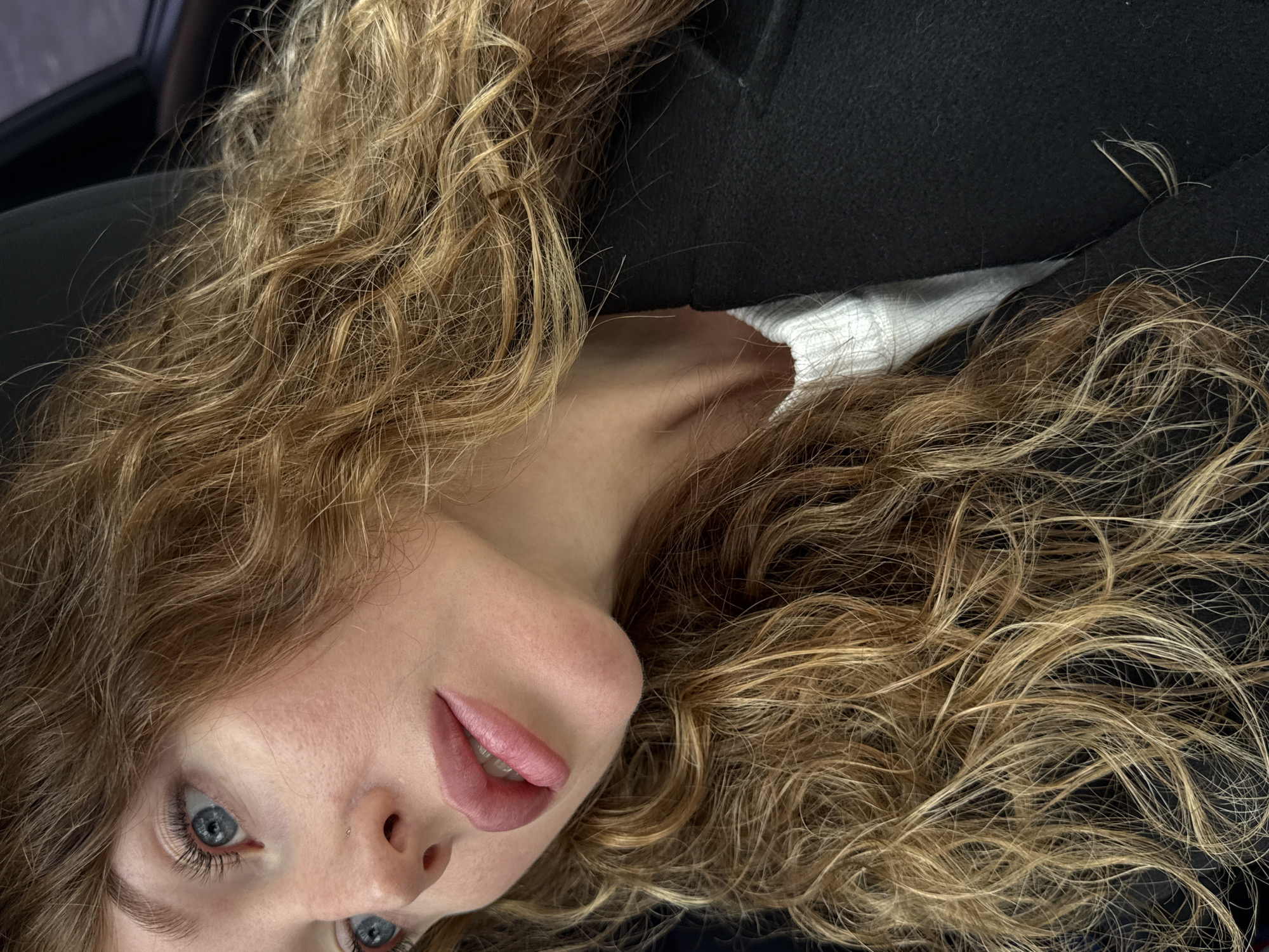 A close-up, slightly rotated image depicts a woman with long, curly, honey-blonde hair that obscures much of the background. Her face is tilted towards the camera, showcasing her pale skin, light blue eyes, and pink lips with the tip of her tongue slightly visible. A black garment is visible behind her neck and to the right, with a glimpse of white fabric between the black garment and her skin. The image's angle and composition suggest it was taken from an overhead perspective. The information about Chaikovsky, Russia is not visible in the image.