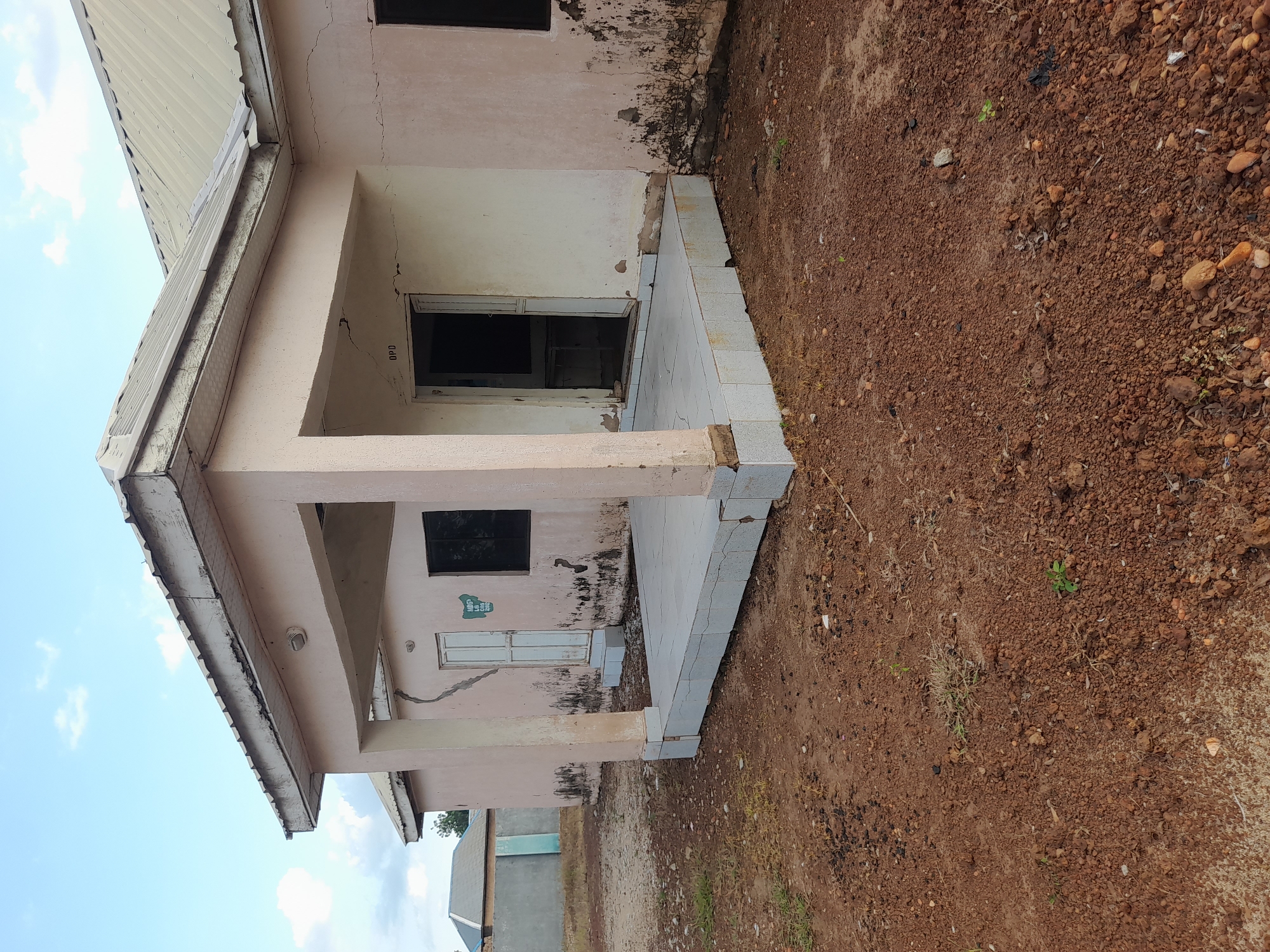 This is a medium-distance, eye-level, outdoor, full-shot photograph of a light pink, single-story building in the city of Ibi, Nigeria. The building has a covered porch with a tiled floor and a white, metal roof with visible supports. The building's exterior walls are weathered and show signs of cracking and discoloration, particularly near the base. There is an open doorway to the right of the porch, marked with the letters "OPD" above it. To the left of the porch, there is a white door with a sign on it. A black-framed window is visible to the right of the open doorway. The ground in front of the building is covered with reddish-brown soil, small stones, and sparse dry grass. In the background, there is another building with a gray concrete wall and a blue roof, and a clear blue sky with scattered white clouds above.