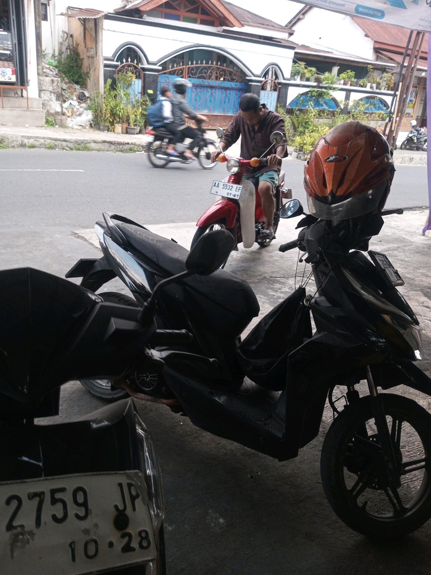 In Mojotengah, Indonesia, two motorcycles are parked on a paved surface in front of a building with a red-tiled roof and white walls. The motorcycle in the foreground is black, with its front wheel visible and a black bag hanging from its handlebars. A license plate on this motorcycle reads "2759 JP 10.28". A red motorcycle is parked to the left, with a license plate that says "AA 5532 EF 07.27". A man in a brown shirt and shorts is riding a red motorcycle in the background, wearing a helmet. Another person, also wearing a helmet, is riding a motorcycle behind him. The street is paved, and there are buildings with arched windows and blue gates visible behind the motorcycles. Green plants are situated in front of these buildings.