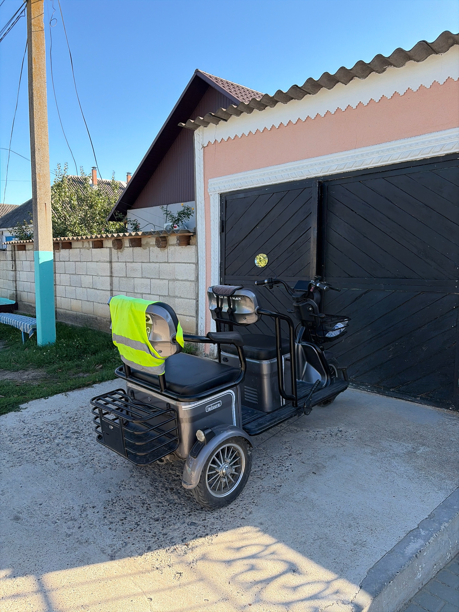 A three-wheeled electric scooter, gray in color with a black seat, is parked on a paved surface in front of a closed black garage door. A bright yellow-green safety vest is draped over the back of the scooter's seat. To the left of the scooter, a section of a light-colored brick wall is visible, topped with wooden beams. Beyond the wall, there is grass, a long wooden bench with a striped cushion, and a concrete pole painted with blue and white stripes. The background shows rooftops of houses and trees under a clear blue sky. The overall scene appears to be in a residential area, possibly in the village of Oxentea, Moldova, given the architectural style and vegetation.