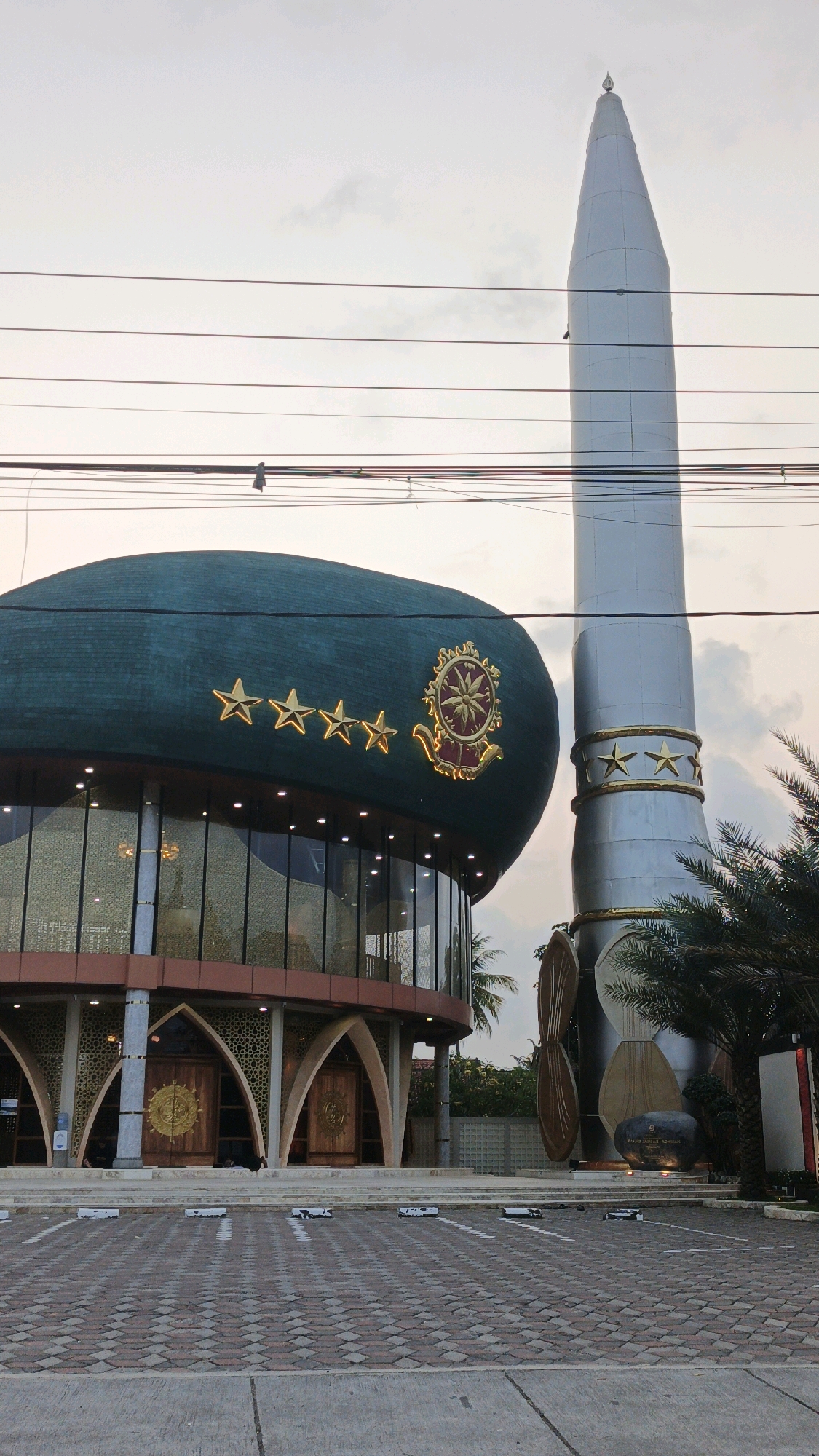 In Desa Cijulang, Indonesia, a striking structure with a large, dark green, beret-shaped dome dominates the foreground. The dome is adorned with five golden stars and a circular emblem in gold and red. Below the dome, the building features large glass windows and arched doorways with ornate wooden doors. To the right of the dome is a tall, silver tower, resembling a rocket, also decorated with golden stars. Palm trees are visible on the right side of the frame. The ground in front of the building is paved with interlocking bricks and concrete. Overhead, power lines crisscross the sky, which is overcast with a pale, hazy light, suggesting either dawn or dusk. A few individuals are seated near the arched doorways.