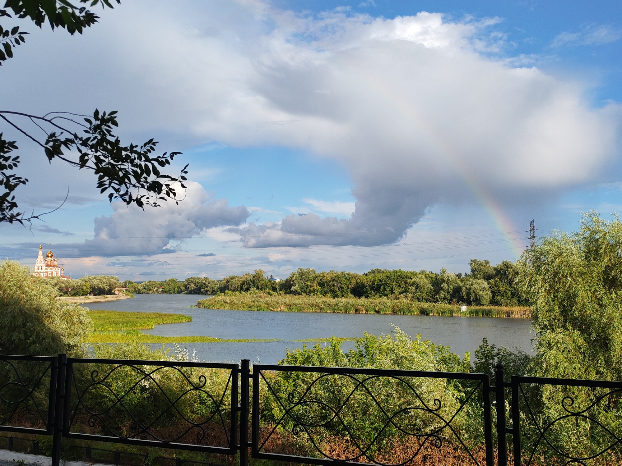 The image depicts a serene view of the Tobol River in Kurgan, Russia. In the foreground, a black wrought-iron fence with decorative patterns runs horizontally across the frame, separating the viewer from the natural landscape. Beyond the fence, lush green trees and shrubs dominate the right side of the image, with a smaller cluster of trees on the left.
The river itself is calm and reflects the blue sky and scattered clouds. Along the far bank, a dense line of reeds and trees creates a natural barrier, with a picturesque church featuring golden domes visible on the far left. A faint rainbow arcs across the sky above a large, billowy cloud formation, suggesting recent rainfall. In the distance, a power line tower stands tall, a subtle reminder of modern infrastructure within the natural setting. The overall atmosphere is peaceful and idyllic, capturing a moment of natural beauty in an urban environment.