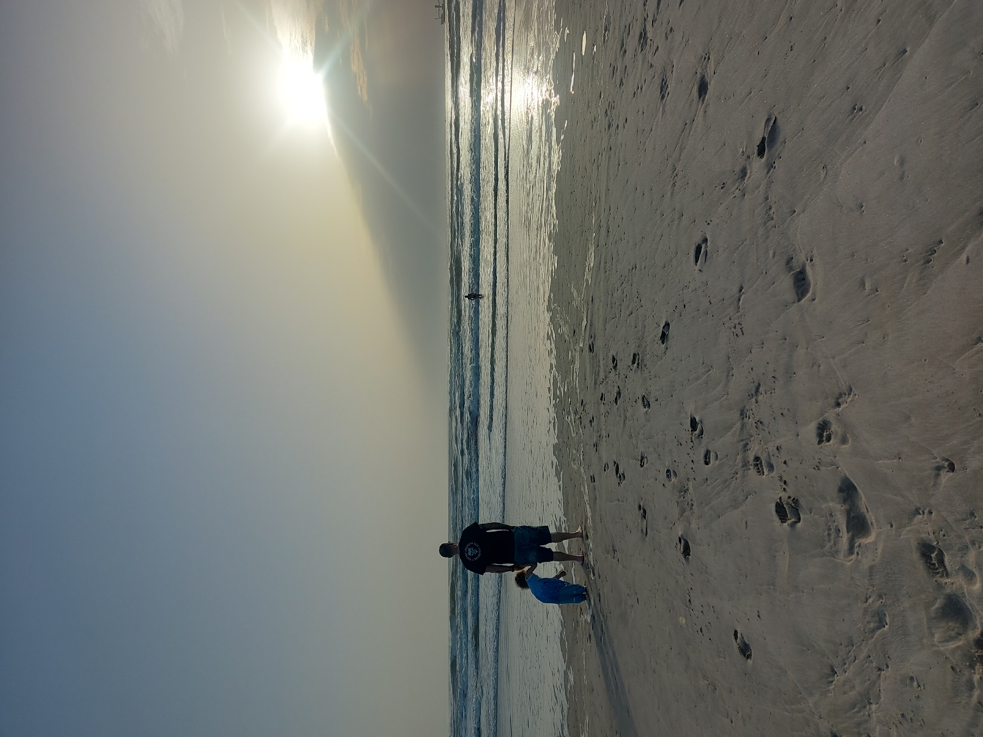 The image captures a beach scene in Figueira da Foz, Portugal. In the foreground, an adult and a child stand at the shoreline. The adult, seen from the back, wears a dark t-shirt and shorts. The child, slightly ahead of the adult, is dressed in a light blue outfit. Footprints are visible in the sand. The ocean extends into the distance, reflecting sunlight. A lone person stands in the water further in the distance. The sky is bright, with the sun near the top of the frame.