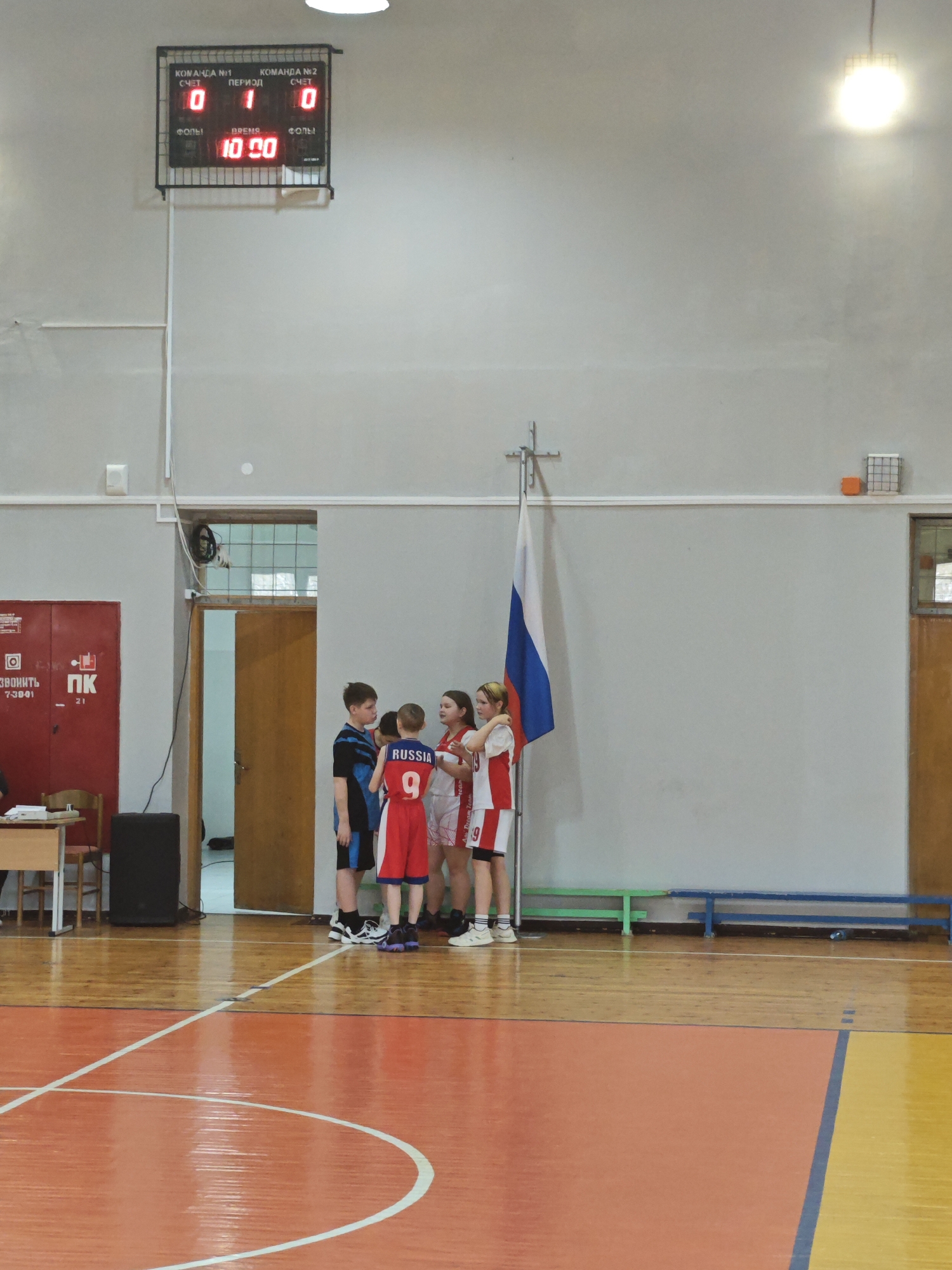 In the town of Markovskii, Russia, a group of young athletes gathers inside a gymnasium. Five individuals, dressed in basketball attire, stand in a huddle near a Russian flag mounted on a stand. One player's jersey displays the word "RUSSIA" along with the number "9." A scoreboard hangs on the wall above them, displaying scores of 0 for both teams, with the time reading "10:20". The gymnasium is a typical indoor sports facility, with wooden floors and a red door with a fire suppression system. Benches line the wall near the flag. The environment suggests a basketball practice or game, possibly at a youth level.