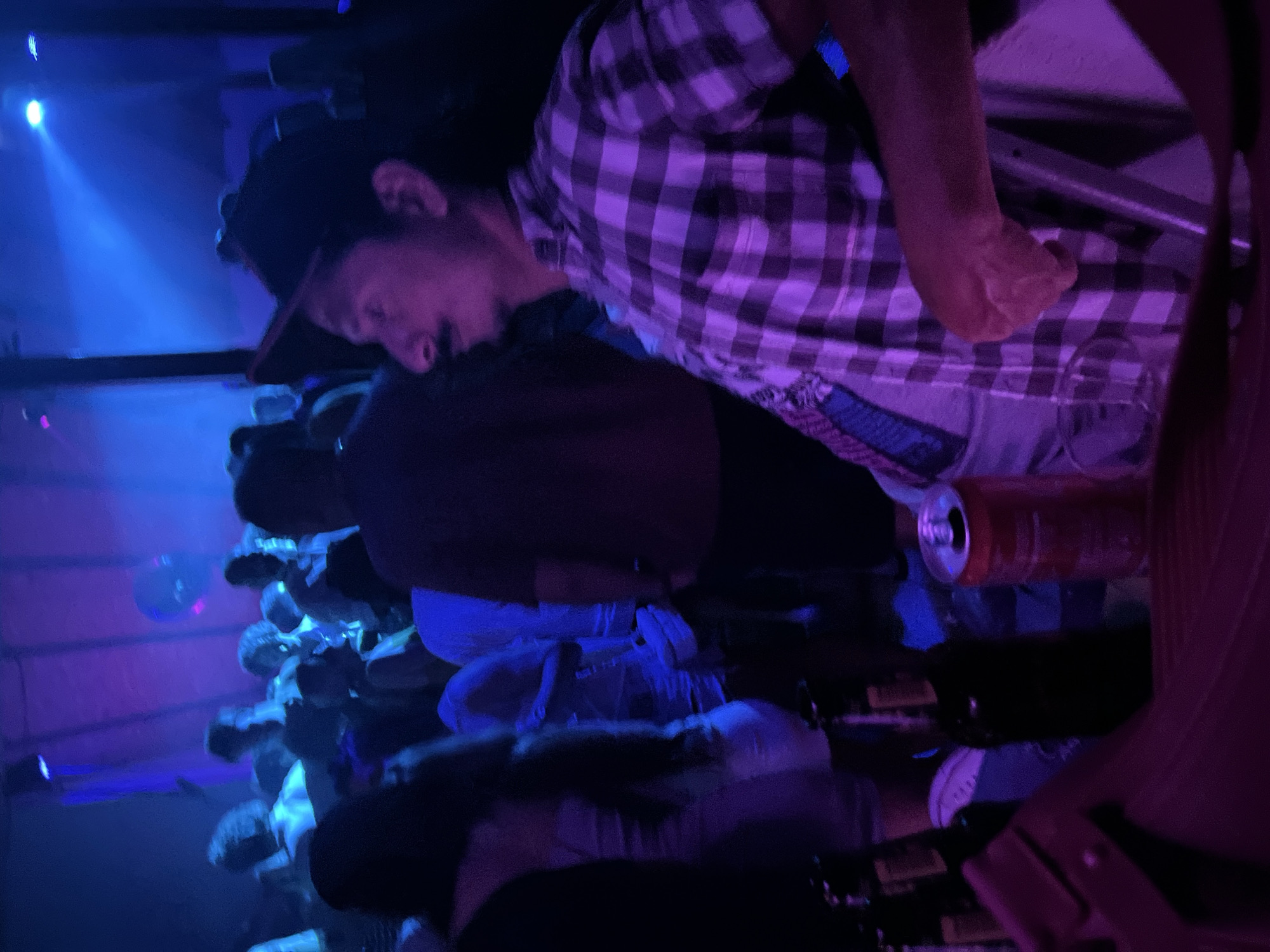 The image depicts a crowded indoor scene, likely a nightclub or bar, illuminated by blue and purple lights. In the foreground, a man wearing a checkered shirt and a cap is visible, looking to the right. His facial features, including a mustache, are discernible. Behind him, other individuals are present, some appearing to be dancing or socializing. Bottles and a can of soda are on a surface in the lower part of the frame. The environment suggests a lively atmosphere with people gathered together. The specific location in Herceg Novi, Montenegro, cannot be determined from the visual information alone.