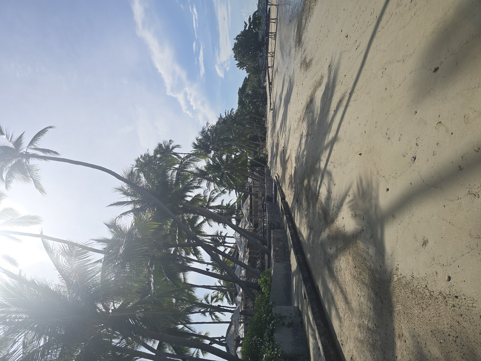 The image depicts a serene beach scene in Baan Chaweng Noi, Thailand, characterized by a row of leaning palm trees casting long shadows on the sandy shore. The sand is a light beige color, with subtle footprints and small pebbles scattered across its surface. To the left, a stone retaining wall marks the boundary between the beach and a vegetated area with chairs and umbrellas, suggesting a resort or beachfront establishment. Beyond the palms, a wooden pier extends into the calm, shallow water. The sky is a soft blue, dotted with wispy white clouds, and the bright sunlight creates a lens flare effect at the top of the image. There are no people visible in the frame.