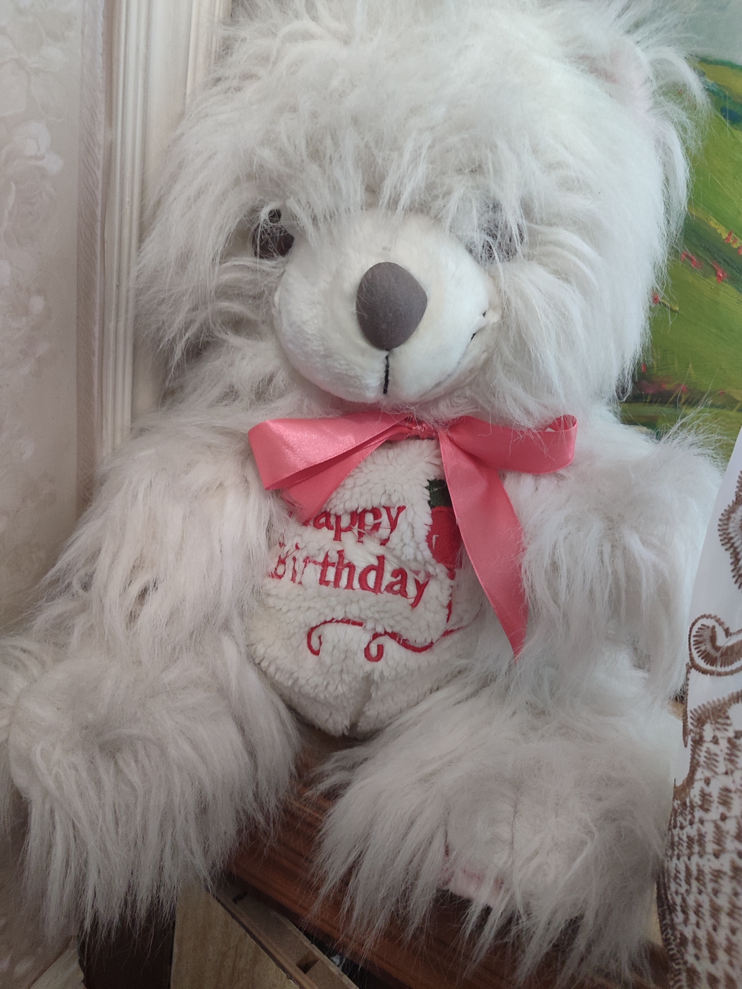 This is a close-up, vertical, indoor shot of a fluffy white teddy bear. The bear is the sole subject, positioned centrally against a neutral, textured background that appears to be a wall with vertical paneling and patterned wallpaper on the left. On the right, a hint of a colorful painting with green and red hues is visible.
The teddy bear has a round, gray nose and dark eyes, partially obscured by its shaggy fur. Around its neck is a bright pink satin bow. Embroidered on its chest in red thread is the text "happy birthday" accompanied by a strawberry graphic. The bear's fur is long and wispy, giving it a somewhat disheveled yet soft appearance. Its arms are resting on its lap, and its legs are mostly out of frame.
The lighting suggests an indoor environment, possibly a room in a home or a shop. There are no indications of time of day or weather conditions. The overall emotion conveyed is one of warmth and celebration, typical of a birthday gift. The embroidery on the bear's chest explicitly points to a birthday event. No other people are visible in the image. The location context of Tashkent, Uzbekistan is not visually supported by any specific elements within the image itself.