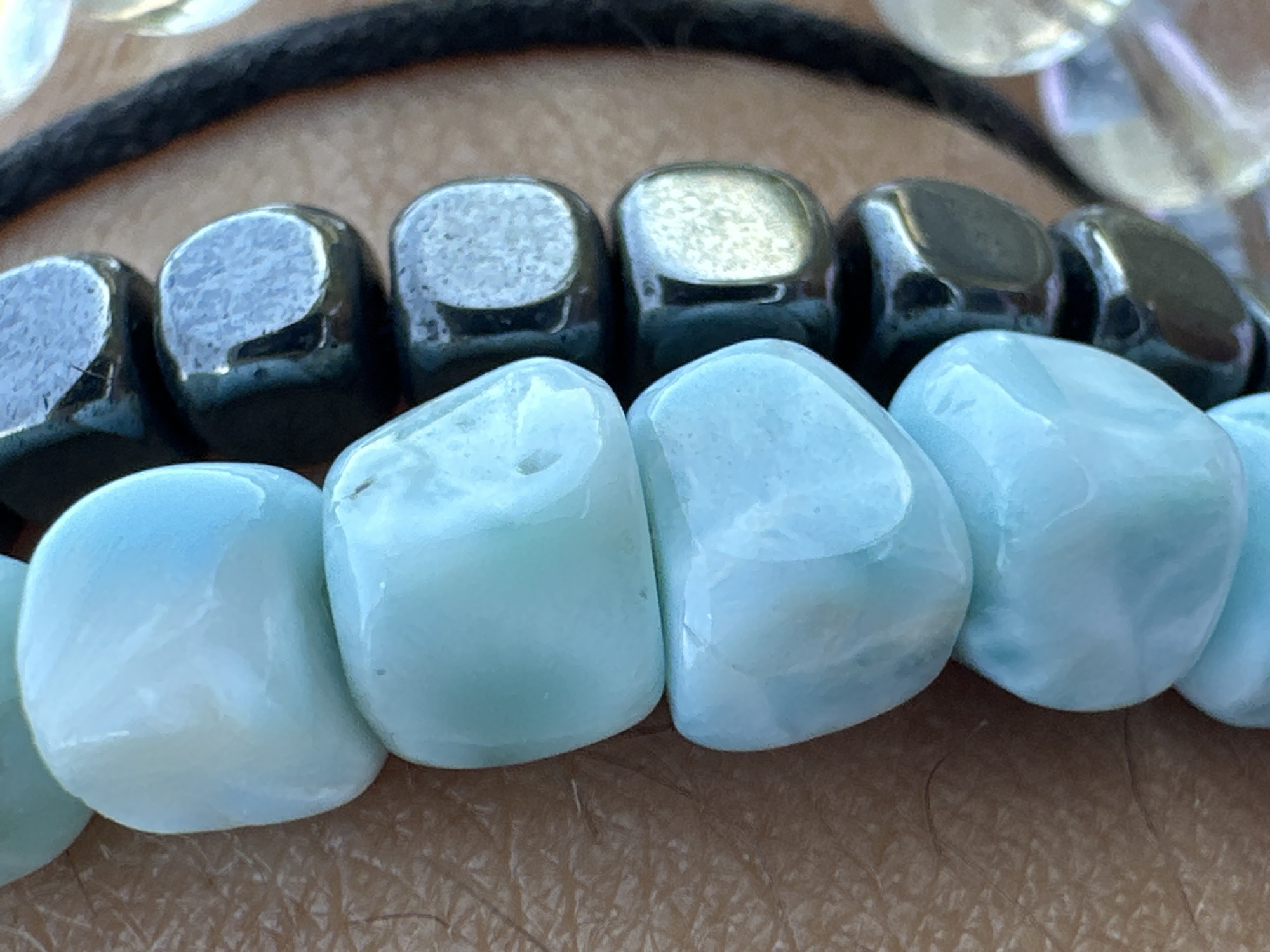 The image shows a close-up of two bracelets. The top bracelet is composed of metallic, cube-shaped beads, black in color, strung together. The bottom bracelet consists of light blue, rounded, irregular-shaped beads. Both bracelets are lying on human skin. A black cord is visible above the top bracelet. Other translucent beads are partially visible at the top of the image. The location is Unknown, Unknown.