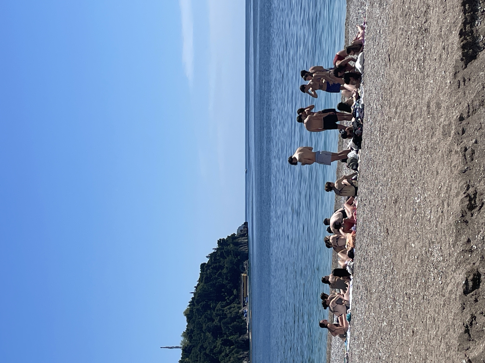 The image shows a beach scene in Budva, Montenegro on a clear, sunny day. A pebble beach is populated with numerous people sunbathing and swimming. A forested hill rises in the background. The water is calm, with a light blue hue. The sky is a cloudless, vibrant blue. People are seen standing and interacting near the water's edge.