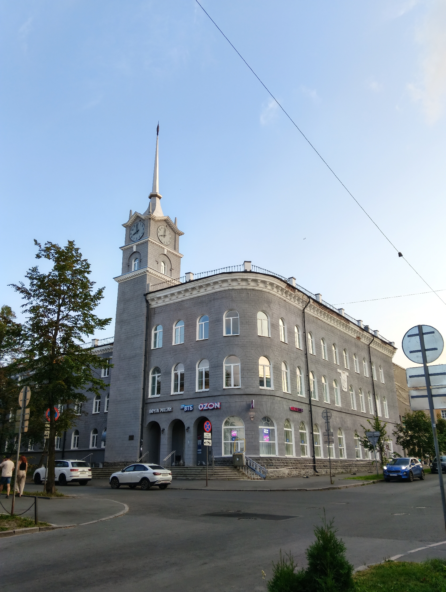 A medium-wide shot captures a large, grey, multi-story building in Petrozavodsk, Russia, with a prominent clock tower. The building exhibits a classical architectural style with arched windows and decorative cornices. Several cars are parked or driving on the street in front of the building. Two pedestrians are walking on the sidewalk. A large tree stands to the left of the building. The sky is clear and blue with a few wispy clouds. Power lines are visible overhead. The scene appears to be set in a downtown area.