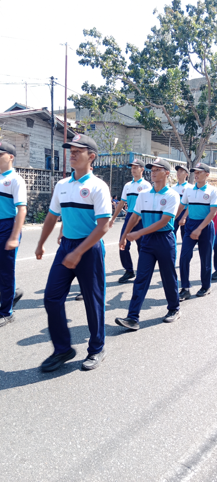 The image shows a group of young men, likely students or members of an organization, marching in formation on a paved road in Tanjung Pinang, Indonesia. They are wearing matching uniforms consisting of blue pants and white and light blue polo shirts with dark blue trim. Each person wears a black baseball cap. The uniforms have a circular emblem on the left chest. The men are walking in a line, and their expressions suggest focus and discipline.
The background reveals a typical urban street scene in Tanjung Pinang, with buildings, trees, and overhead utility wires visible. One building, partially seen on the left, appears to be a single-story structure with a pitched roof and light-colored walls. Another building in the background is taller and made of concrete. Trees with green foliage line the street, casting shadows on the road, indicating it is daytime and sunny. The road surface is asphalt, with some visible cracks and texture. The overall impression is of a formal procession or parade taking place in a residential or commercial area of the city.