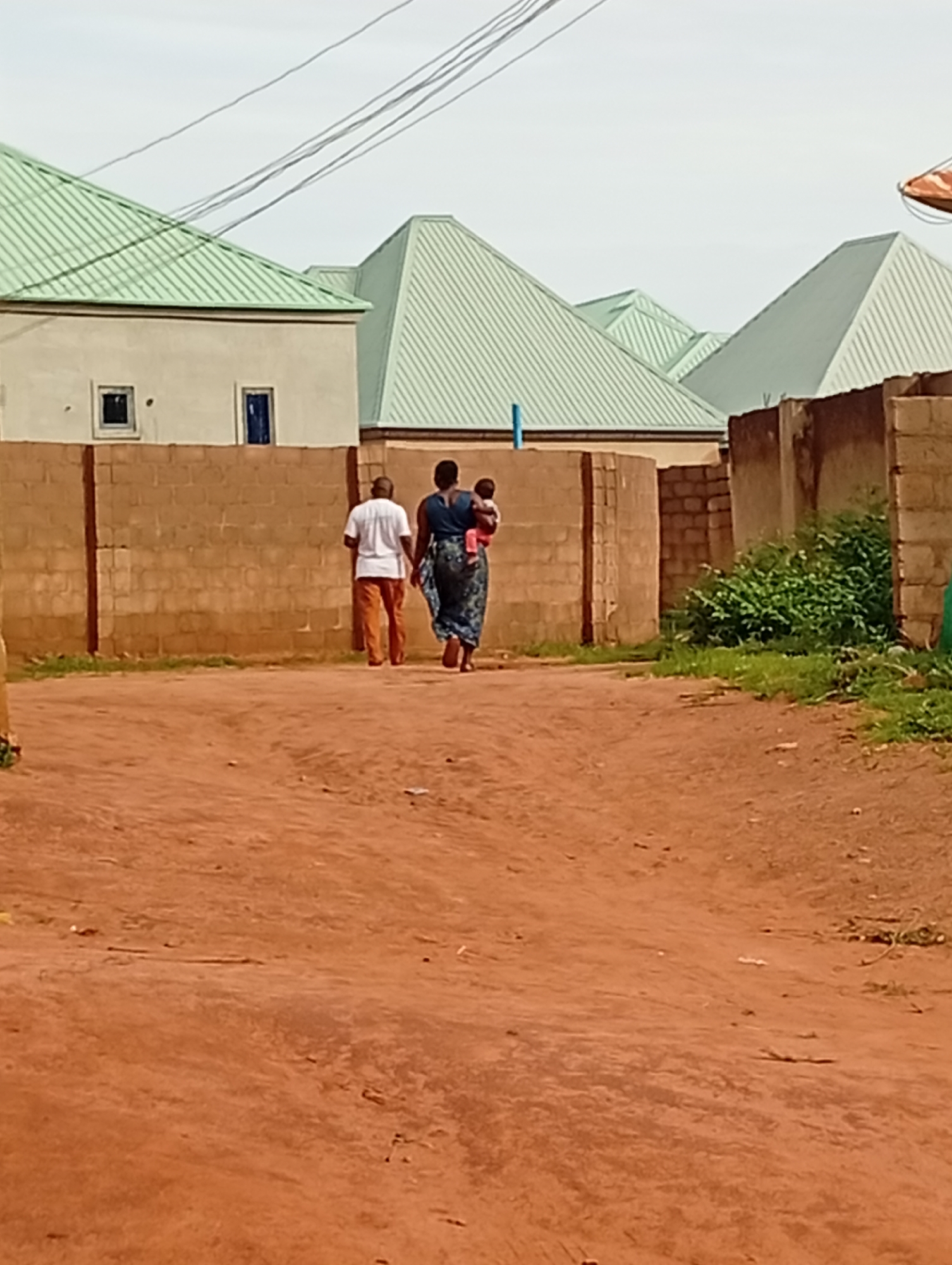 The image depicts an outdoor scene on a dirt road in Tumfure, Nigeria. Two individuals, a man and a woman carrying a baby, are walking away from the camera towards the right side of the frame. The man is wearing a white t-shirt and orange pants. The woman is wearing a dark blue dress with a patterned skirt and is holding a baby wrapped in a pink cloth. Behind them, a low brick wall runs horizontally across the middle ground. Beyond the wall, several buildings with distinctive green, corrugated metal roofs are visible. The sky is overcast and light in color. The ground is covered in reddish-brown dirt, with some scattered debris. Vegetation, primarily green bushes, is growing to the right of the brick wall. Overhead, power lines stretch across the upper part of the image.