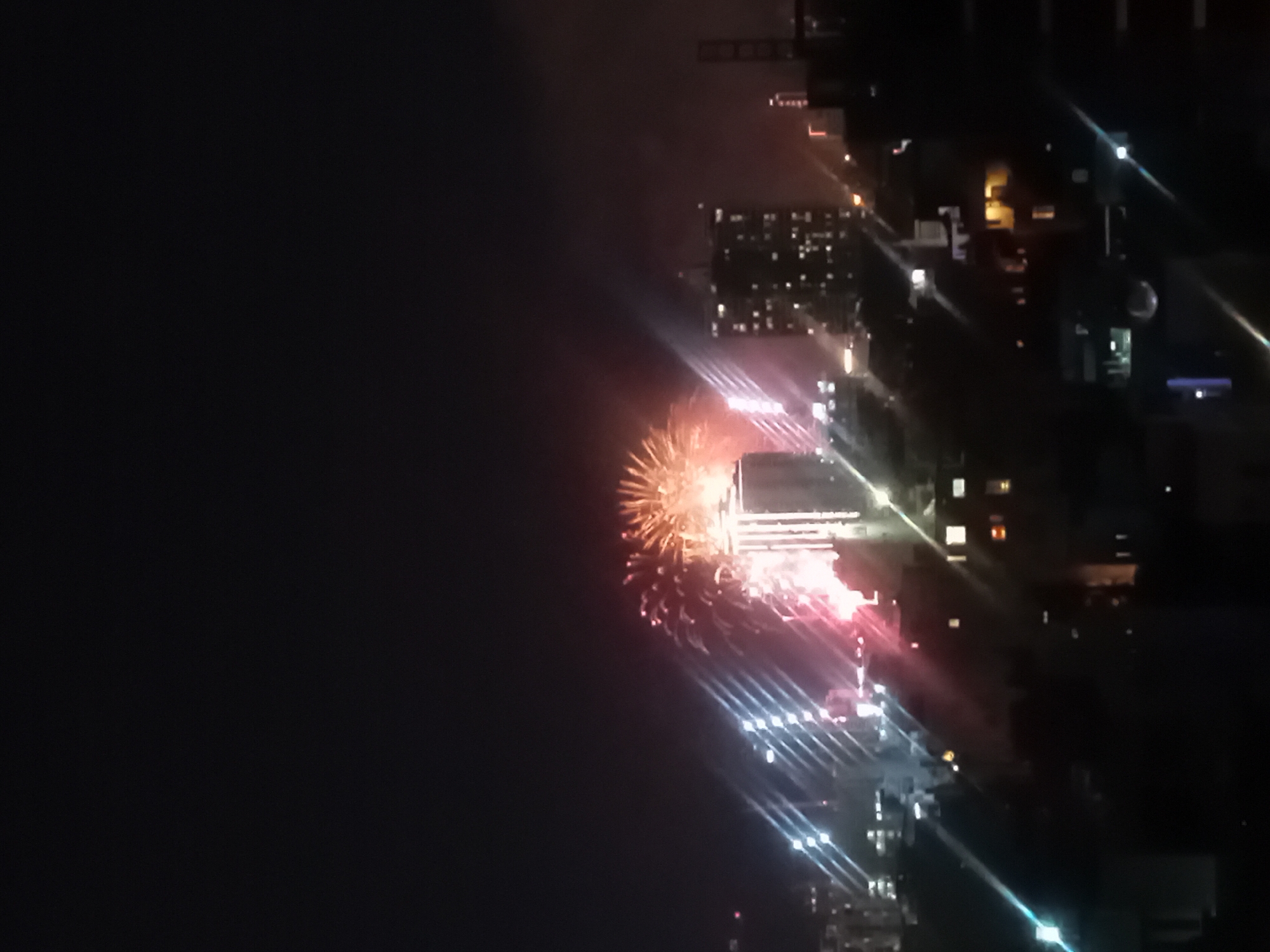 The image is a nighttime scene of a city with fireworks. In the foreground, there are silhouetted buildings and trees. The middle ground features a cluster of illuminated buildings, including a tall, modern skyscraper with a brightly lit facade that appears to be the focal point. There are also other buildings with visible windows and lights. In the background, the sky is dark with numerous bright fireworks exploding in various colors, primarily orange, yellow, and pink. Beams of light, possibly from searchlights or other illuminated features, cut through the scene, creating streaks of light across the image. The location appears to be the city of Đà Nẵng, Vietnam, given the presence of the Han River bridge, which is not clearly visible but is hinted at by the urban landscape and the possibility of a celebratory event.