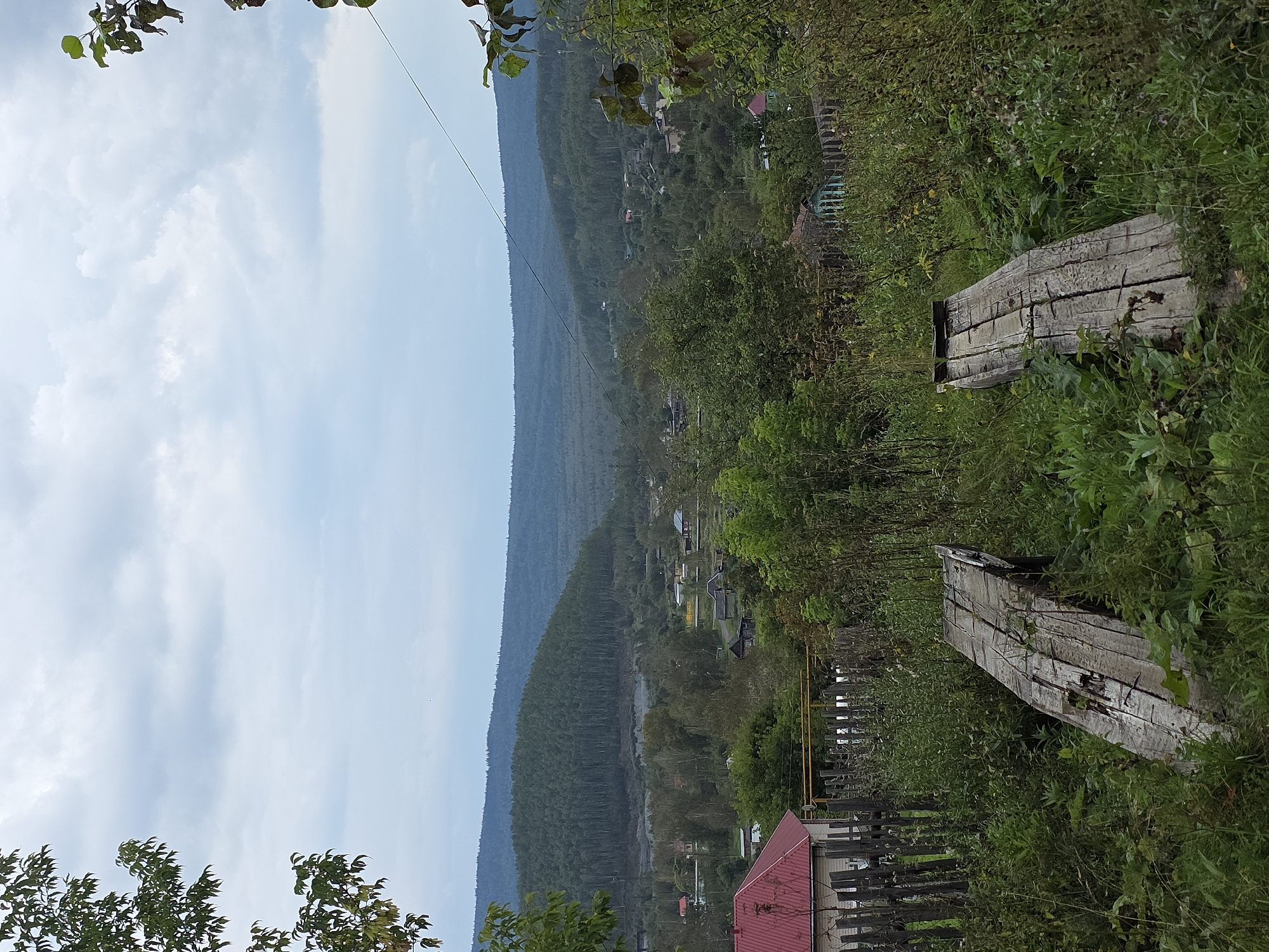 The image shows an outdoor, elevated view of a rural landscape under an overcast sky. In the foreground, two weathered wooden planks are positioned diagonally, partially obscured by green foliage. To the left, a wooden fence and the corner of a house with a red roof are visible. The middle ground reveals a village with scattered houses, surrounded by trees. The background features a densely forested hill receding into a hazy, mountainous horizon. The scene appears to be located in Magnitka, Russia, characterized by its natural surroundings and rustic village setting. There are no people or animals present in the image.
