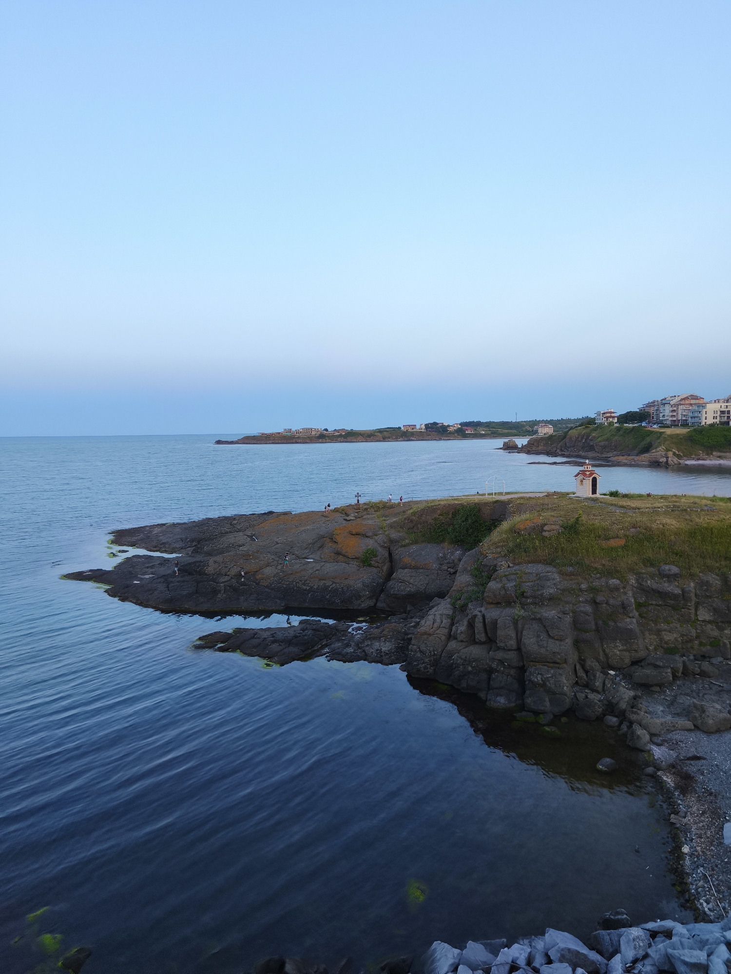 The image captures a serene coastal landscape at dusk, likely within the vicinity of Tsarevo, Bulgaria. The scene is dominated by the vast, calm expanse of the Black Sea, its deep blue waters reflecting the soft hues of the twilight sky. A rocky peninsula juts out into the sea, its rugged surface covered in sparse grass and dotted with a few people. On the easternmost point of this peninsula stands a small, white chapel with a red roof, adorned with a cross.
In the background, the coastline of Tsarevo unfolds, featuring a cluster of multi-story buildings with light-colored facades, suggesting a residential or tourist area. The buildings are nestled on a gentle incline overlooking the bay. The overall atmosphere is peaceful, with the diminishing daylight casting a tranquil glow over the entire scene. The presence of a few individuals on the rocks and near the chapel suggests a leisurely evening stroll or quiet contemplation by the sea.