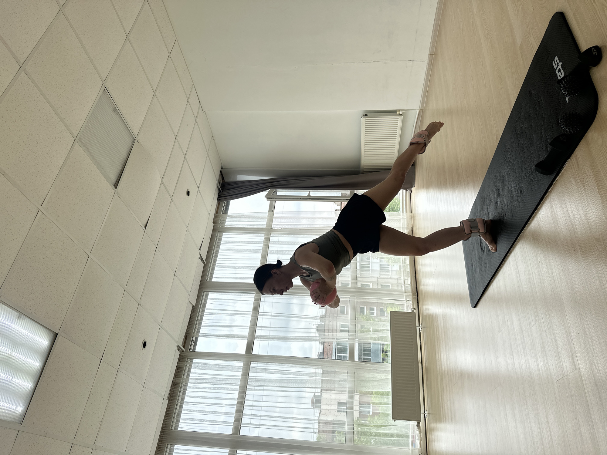 A woman is performing a balancing exercise on a black yoga mat in a bright room. She is wearing a dark green tank top and black shorts, with pink ankle weights on her lower legs. She is holding a pink ball in her hands, clasped in front of her chest, and is balancing on her left leg with her right leg extended behind her. The room has a light-colored wooden floor and is illuminated by overhead fluorescent lighting. Large windows with white sheer curtains line one wall, offering a view of apartment buildings outside. Radiators are visible beneath the windows. The setting appears to be a dance or exercise studio, possibly located in Votkinsk, Russia, given the architectural style of the buildings visible through the windows and the type of radiators, which are common in Russian residential buildings.