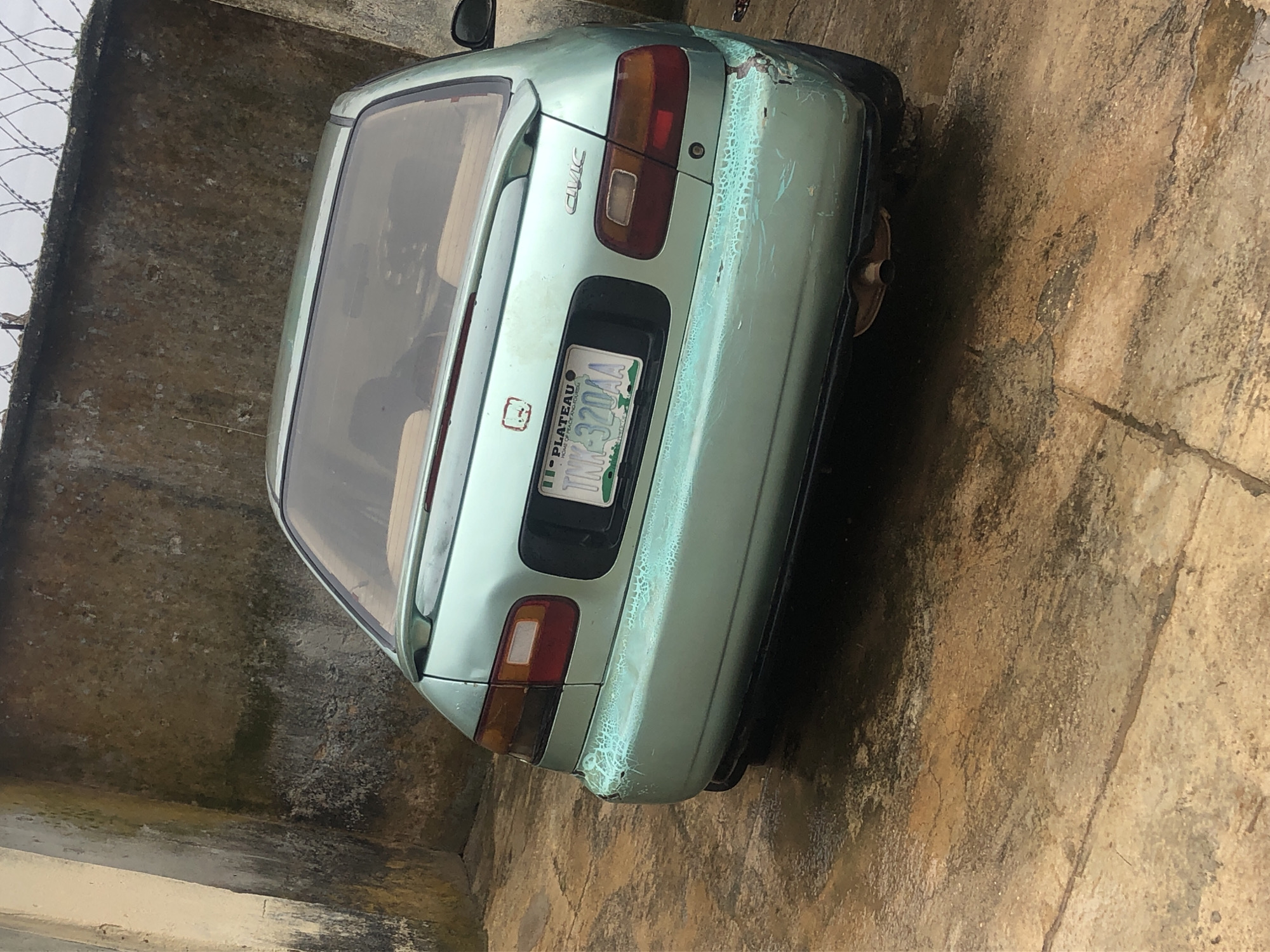 A rear, three-quarter view of a light green Honda Civic sedan is parked on a wet, stained concrete surface. The car has a spoiler on the trunk, a license plate that reads "TNK-320AA", and a "CIVIC" badge on the right side of the trunk. The rear bumper appears damaged and has peeling paint. The car is parked in front of a dirty, textured concrete wall, partially covered in moss. A barbed wire fence runs along the top of the wall. The sky is visible through the fence. The overall scene suggests an outdoor, possibly neglected urban or semi-urban environment.