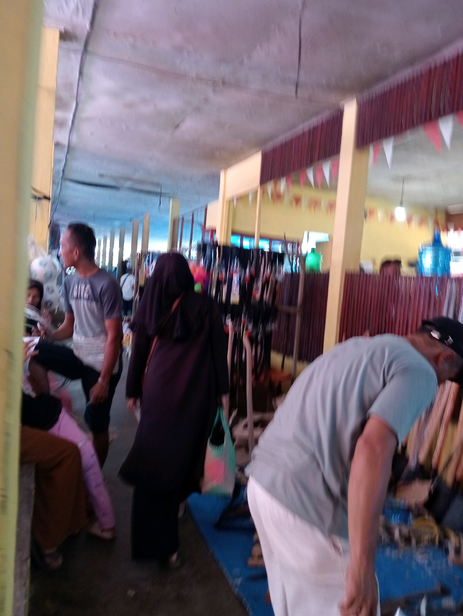 The image depicts a scene at the Taman Cinema in Pariaman, Indonesia, a public space that appears to be hosting an event or market. In the foreground, a man in a light blue t-shirt and white pants is bent over, his back to the camera. To his left, a person wearing a black hijab and dark clothing walks away, carrying a colorful tote bag. Further to the left, a man in a gray t-shirt is interacting with someone out of frame, while a glimpse of other individuals and objects suggests a bustling atmosphere. The environment includes a covered walkway with visible support columns and decorative banners, typical of an outdoor event space. The background shows stalls with items for sale, possibly clothing or accessories, and a large blue water cooler. The overall impression is one of a lively gathering in a communal area.