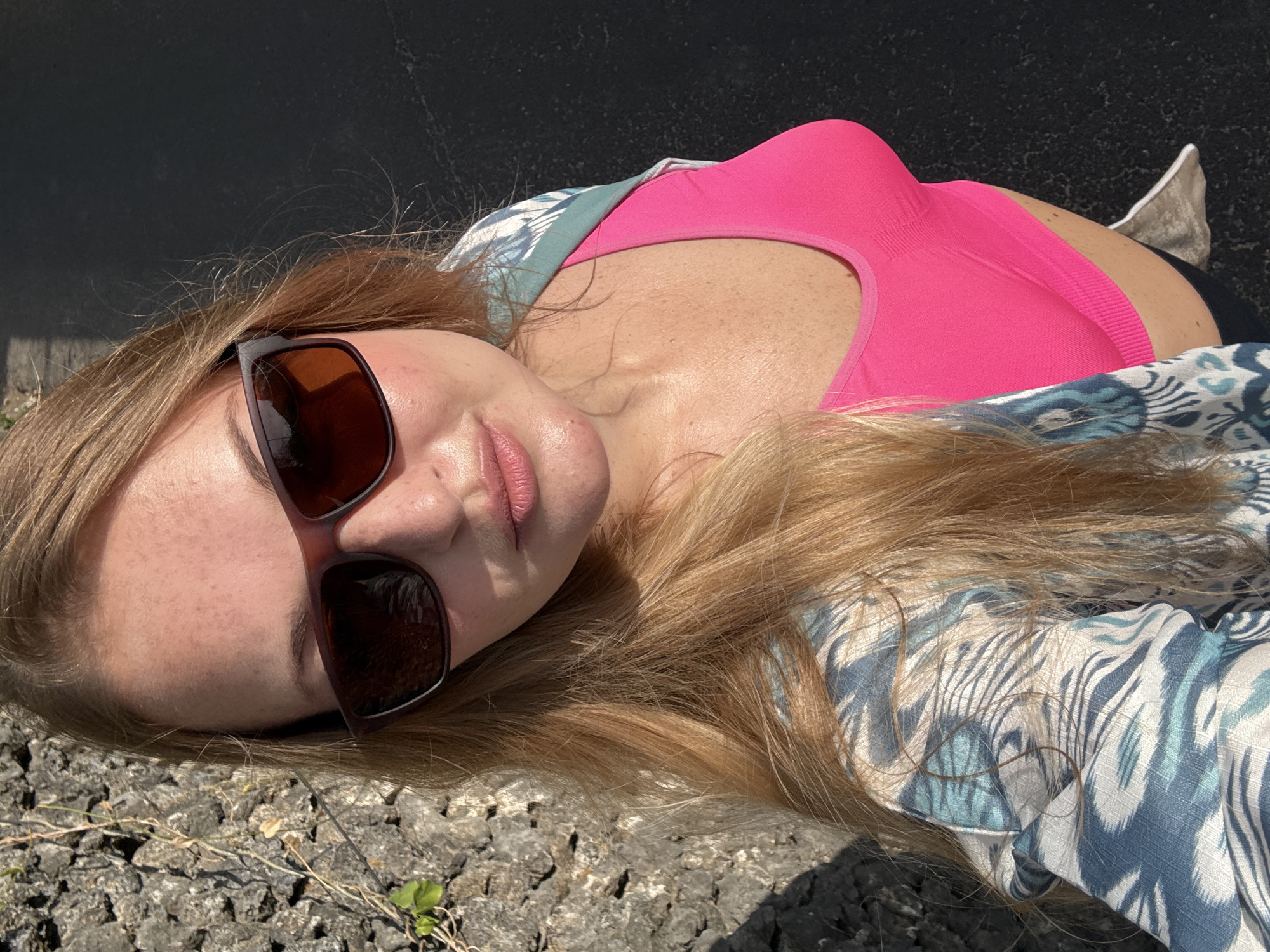 A blonde-haired woman, wearing sunglasses and a bright pink sports bra beneath an open, patterned blue and white shirt, is pictured outdoors. Her face is partially obscured by the sunglasses. She is standing near a rough, grey stone wall on her left and a dark, asphalt-like surface on her right. The lighting suggests it is daytime, with sunlight casting shadows. The environment does not provide any specific visual cues to pinpoint a location within the city of Denpasar, Indonesia.