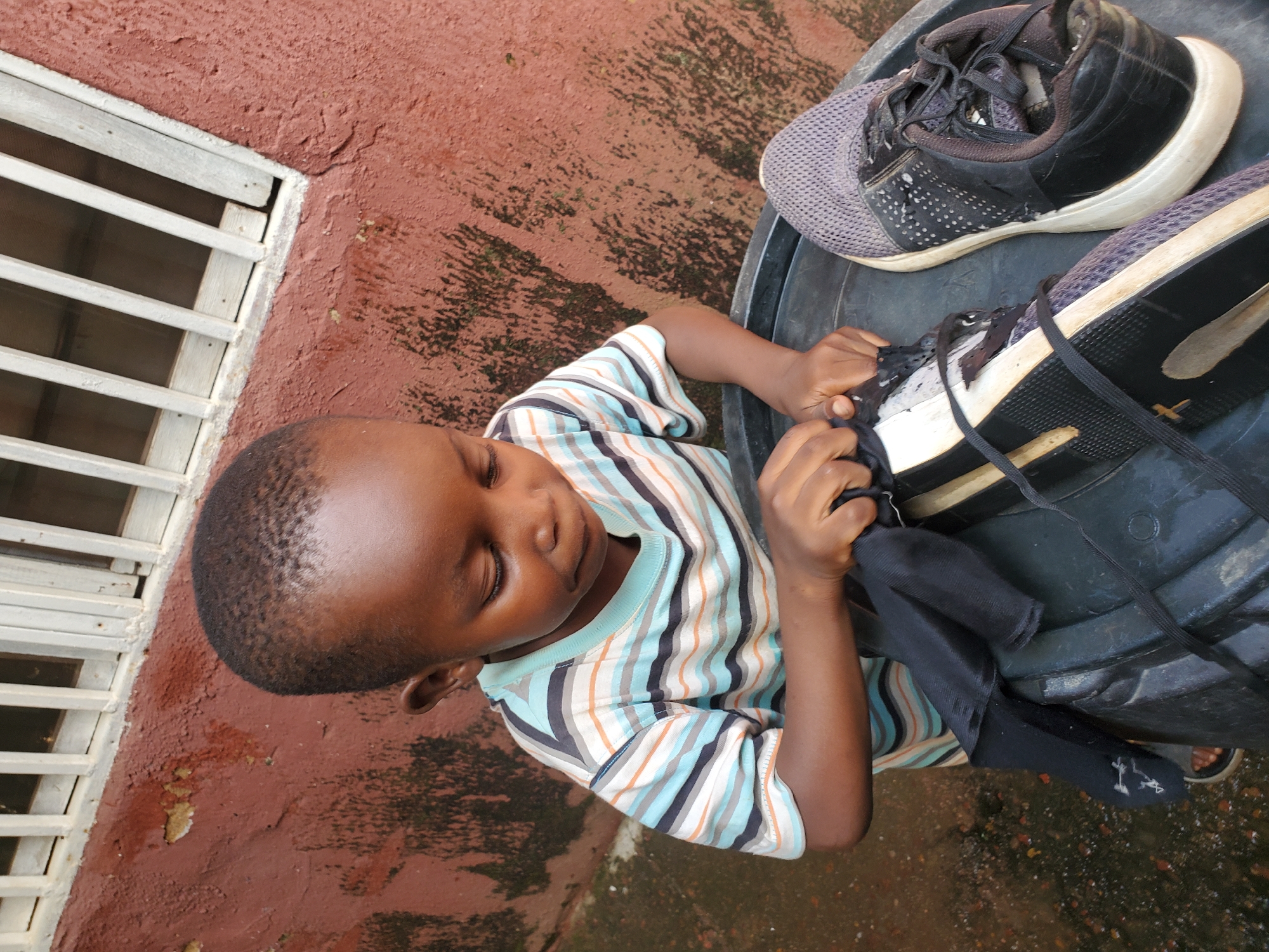 A young boy, appearing to be around five years old, with dark skin and short hair, is shown from a high angle, looking down with a focused expression. He is wearing a horizontally striped t-shirt in shades of blue, grey, black, and orange. His hands are actively engaged in manipulating a black sock, which he is pulling out of or preparing to put into a black plastic container. Inside this container, and partially outside, are two athletic shoes. One shoe, to the left, is black and white with visible wear. The other shoe, to the right, is grey and black, also showing signs of use. The container is resting on a wet, possibly concrete, ground that has scattered debris, suggesting an outdoor environment. In the background, a textured, reddish-brown wall with dark patches of moss or algae is visible, surmounted by a white barred window or opening. The overall scene suggests an activity taking place in a humble outdoor setting, possibly a residential compound or yard in the city of Jos, Nigeria.