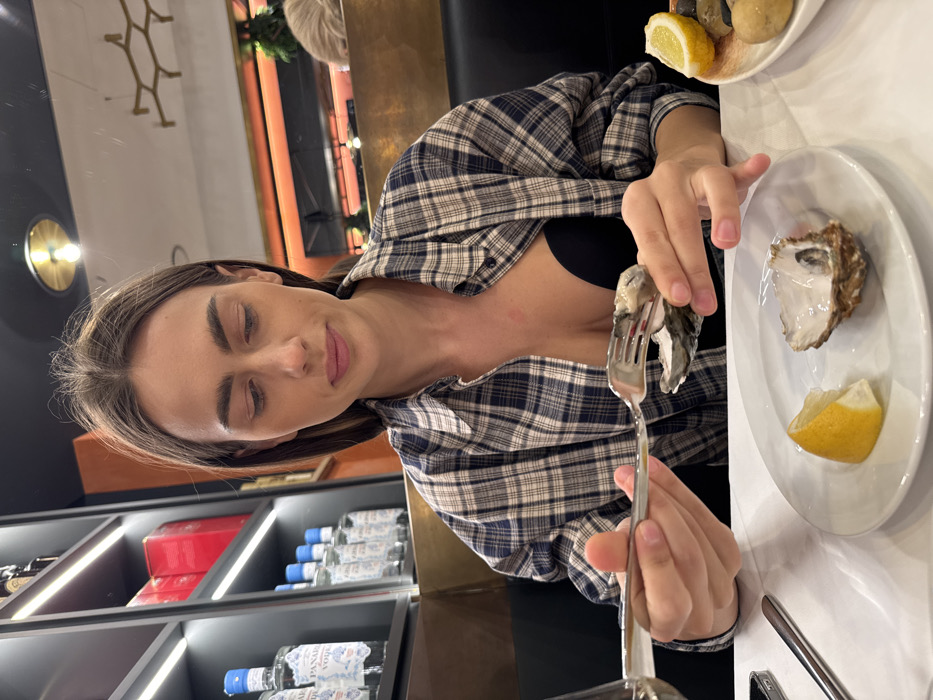 The image presents a candid, mid-shot view of a woman seated at a table in an indoor restaurant setting. While identified as being in Verona, Italy, specific architectural or branding cues within the frame do not allow for the precise identification of the establishment. The woman, characterized by long, dark brown hair, fair skin, and natural makeup, is wearing a blue and white plaid button-up shirt, unbuttoned to reveal a black bra. Her gaze is directed downwards, focusing on the activity before her.
She is actively consuming oysters. Her left hand holds an oyster in its shell, while her right hand manipulates a silver fork, positioned to extract the oyster's contents. On the white tablecloth, a white ceramic plate holds a consumed oyster shell and a lemon wedge. To the right, a partially visible dish contains smooth stones and an additional lemon wedge. A silver smartphone and the stem of a clear glass are also discernible on the table.
The background reveals a modern, dimly lit interior. To the left, dark shelving units with integrated strip lighting display multiple bottles of "PANAREA ISLAND VODKA" and several red boxes. To the right, dark booth seating, an orange-brown wall panel, and a mirror reflecting a circular, golden-rimmed light fixture and the head of another patron are visible. The overall ambiance suggests an evening dining experience.