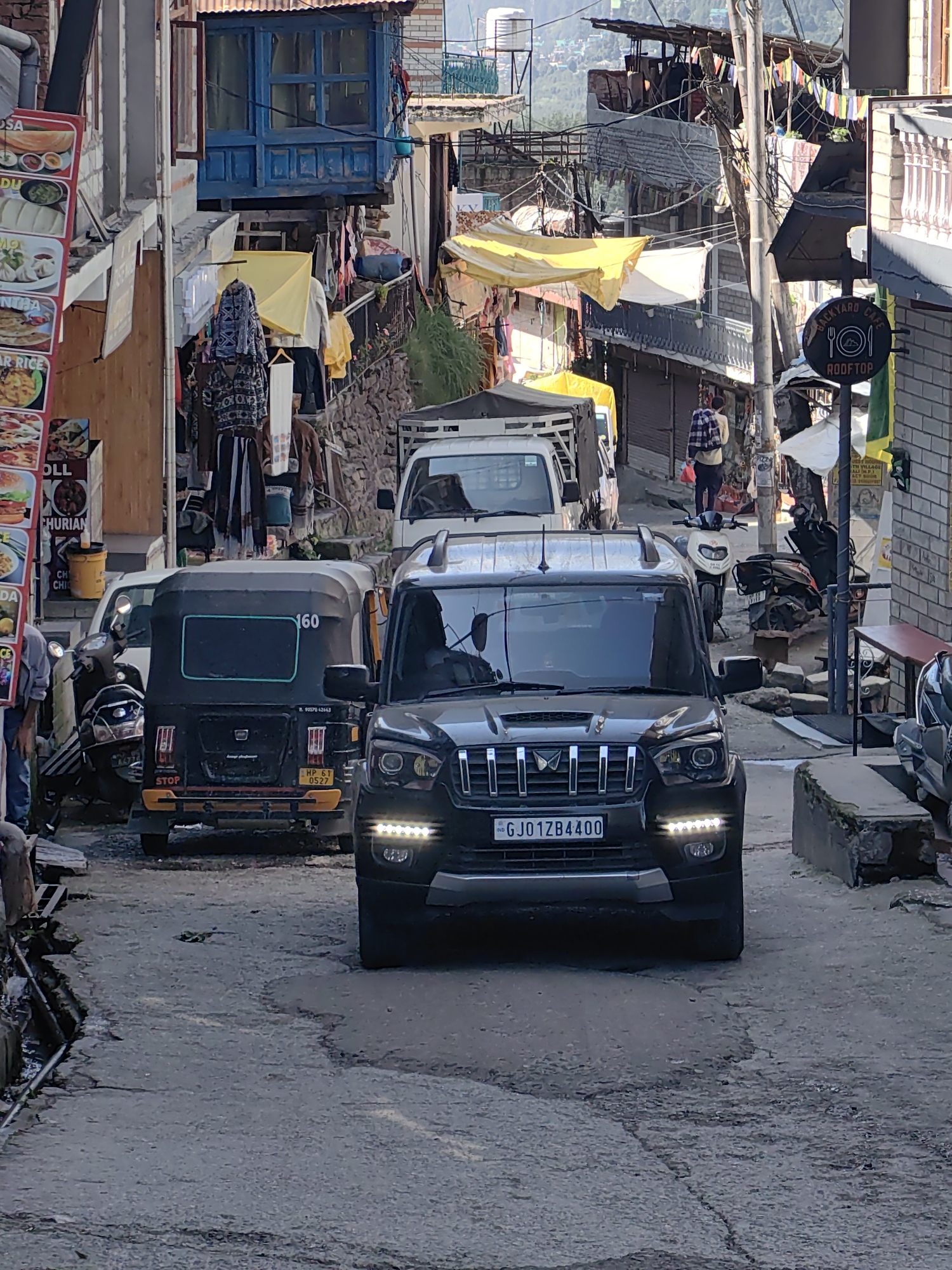 The image depicts a narrow street in Vashisht, India, lined with buildings and shops. A black Mahindra Scorpio SUV with license plate "GJ01ZB4400" is prominently featured in the foreground, driving down the street. To its left is a black and yellow auto-rickshaw with the number "160" visible. Further back, a white truck is parked. Shops on either side of the street display goods and signage, including a menu board with images of food items. Clothing items hang outside some establishments. A person in a plaid shirt and dark pants walks away from the camera further down the street. The street is paved with asphalt, showing some cracks and wear. The architecture of the buildings is a mix of stone and plaster, with some balconies and colorful window frames. Yellow tarpaulin sheets are visible over some structures. Electrical poles and wires are present throughout the scene. The overall impression is of a bustling, lived-in street in a mountainous region.
