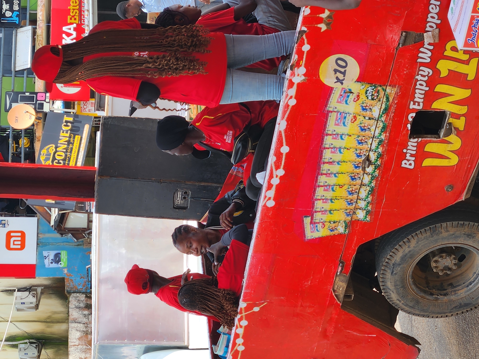 In an outdoor urban environment, possibly a street market or roadside area, a bright red, open-top vehicle resembling a modified truck or cart is parked. The vehicle is decorated with images of packaged snacks and the text "Bring 10 Empty Wrappers WIN 1R". A group of people are interacting within and around the vehicle.
In the foreground, a woman with long, braided hair, wearing a red t-shirt and a red baseball cap, stands facing away from the camera. She is holding a phone. To her left, two other women are seated inside the red vehicle. One woman, wearing a black headscarf and a red t-shirt, is looking to her left. The other woman, in a black jacket and red t-shirt, is facing the woman with braids and appears to be speaking to her.
In the background, several signs are visible, including one with the "MI" logo and another advertising "CONNECT". The overall scene suggests a promotional event or a mobile sales operation. The lighting indicates daytime.