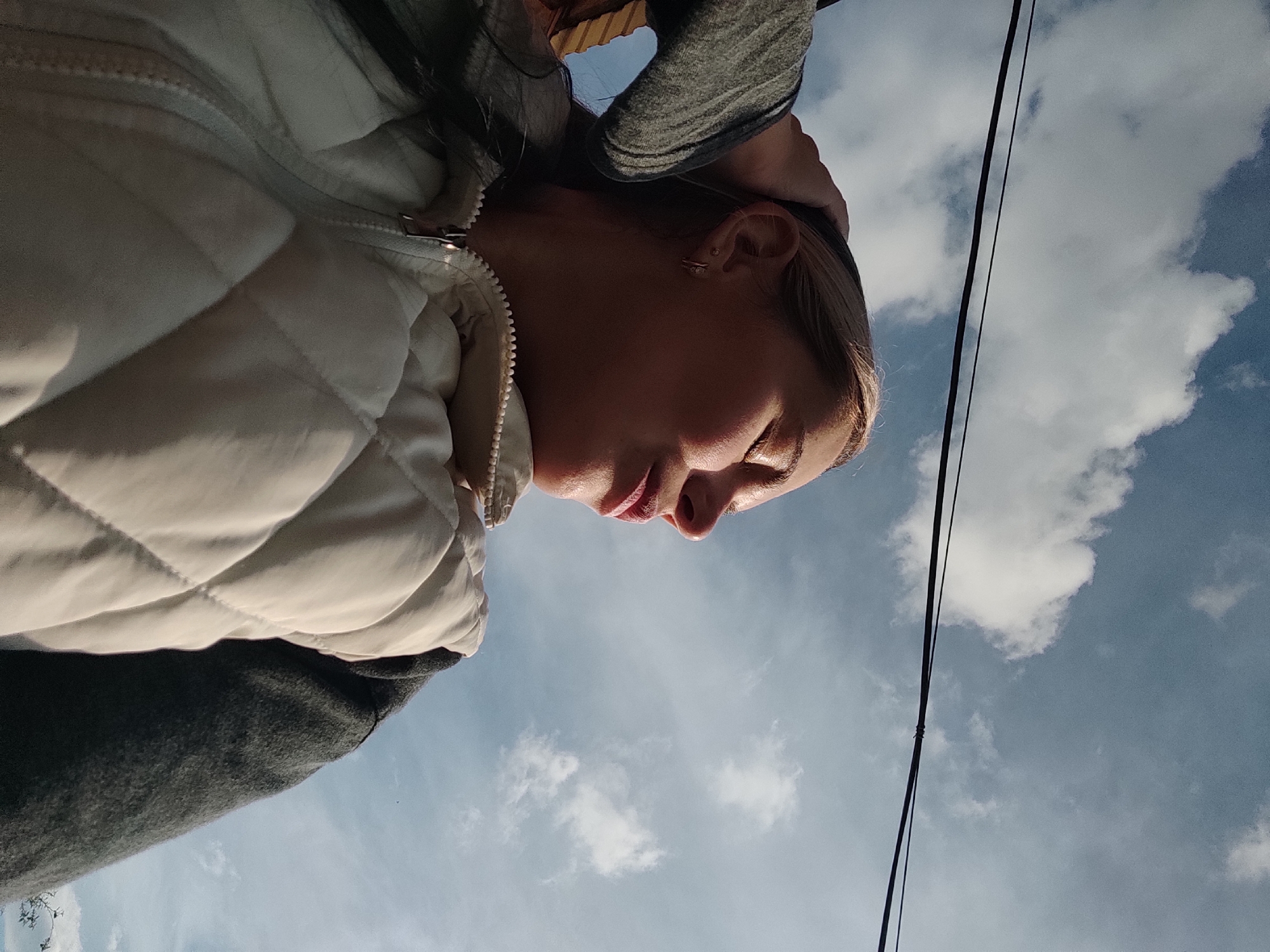 A woman is photographed from a low angle, gazing downward. She wears a light-colored, quilted jacket over a gray garment. Her hair is blonde. Above her, the sky is visible with scattered clouds and two parallel, vertical black electrical wires. The location is identified as Ольховка, Russia.