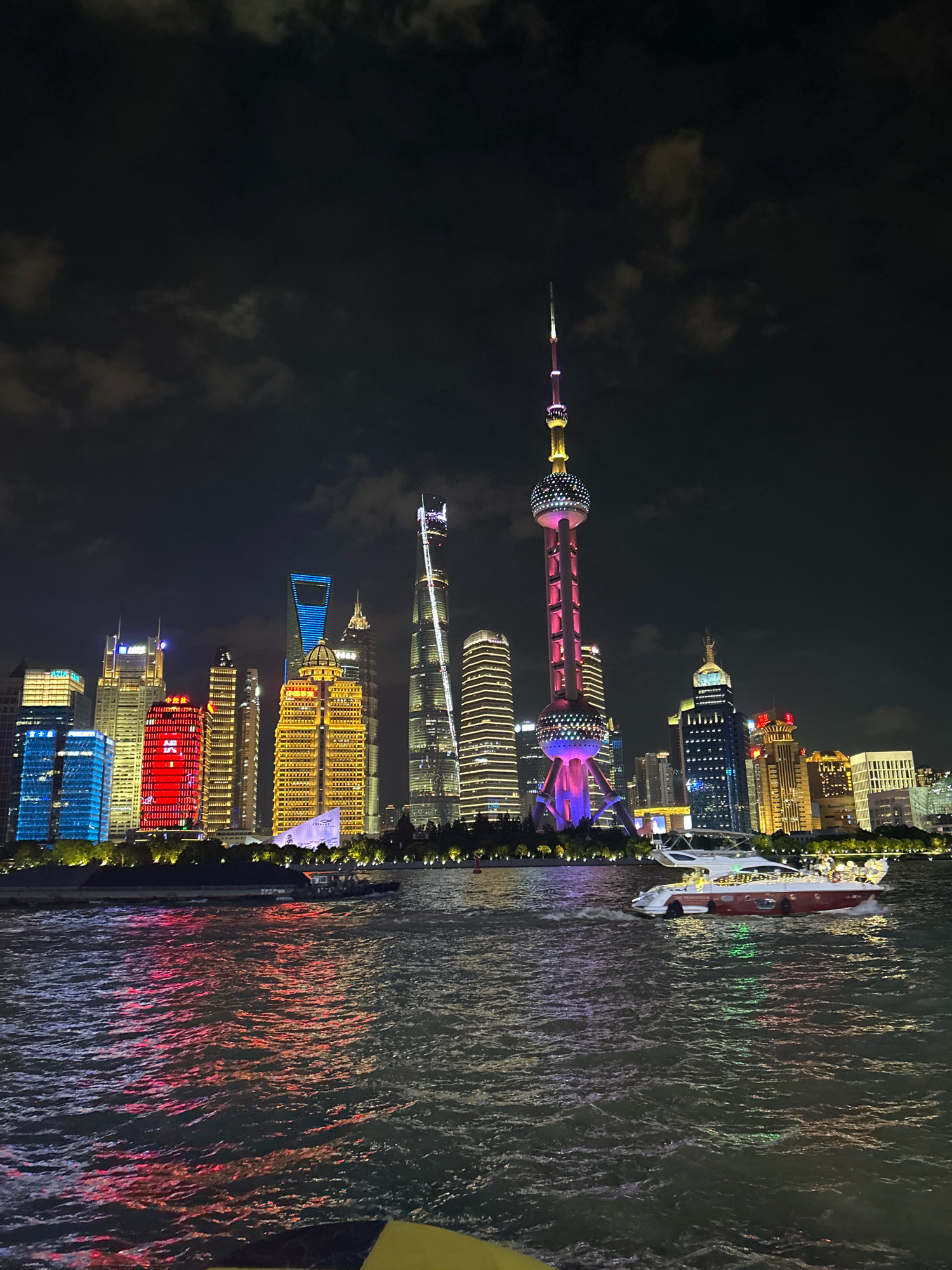 An nighttime exterior shot captures the illuminated skyline of Shanghai's Pudong district, viewed from across the Huangpu River, likely in the Hongkou District. The water in the foreground reflects the colorful lights of the buildings, which are a mix of modern skyscrapers and the iconic Oriental Pearl Tower. Several boats are visible on the river, with one luxury yacht in the middle ground featuring red and white detailing and illuminated windows. The sky is dark with some cloudy texture.