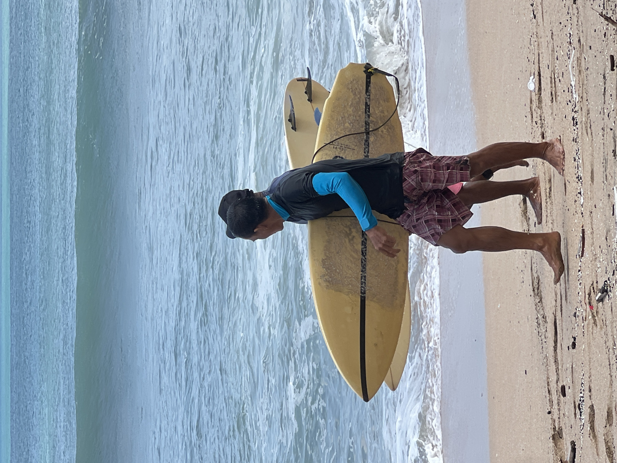 Two men are walking on the beach in Kuta, Indonesia, carrying two surfboards. The man in the foreground is wearing a black long-sleeved shirt with blue sleeves, and plaid shorts. He is carrying a yellow surfboard. The man behind him is wearing a dark shirt and is also carrying a surfboard. The ocean has moderate waves, and the sky is overcast. The beach is sandy and strewn with debris.