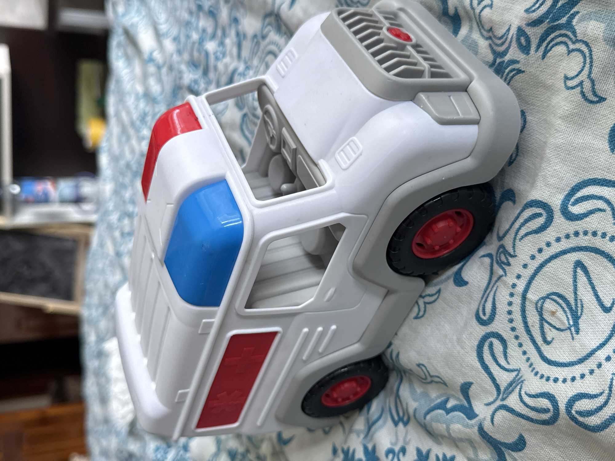 The media file features a close-up, angled shot of a white toy ambulance with red and blue accents, resting on a blue and white patterned fabric. The toy ambulance is the primary object, and it appears to be a child's plaything. The fabric it rests on is likely a blanket or bedsheet with an intricate, repeating pattern of swirling blue designs.
In the background, out of focus, is a dark wooden surface with some objects on it, including what appears to be a shelf or frame. There are also hints of other items, possibly books or toys, in the blurred background. The lighting suggests it is daytime, with natural light illuminating the scene. No people are visible in the frame.
The toy ambulance is detailed, with visible wheels (black with red rims), a steering wheel and dashboard inside the cab, and a grill on the roof. Red and blue lights are present on the top of the cabin, and red rectangular markings are on the side. The overall impression is a domestic setting, possibly a bedroom or playroom. The image is static, with no activity taking place beyond the object being placed on the fabric. There is no visible text in the image.
