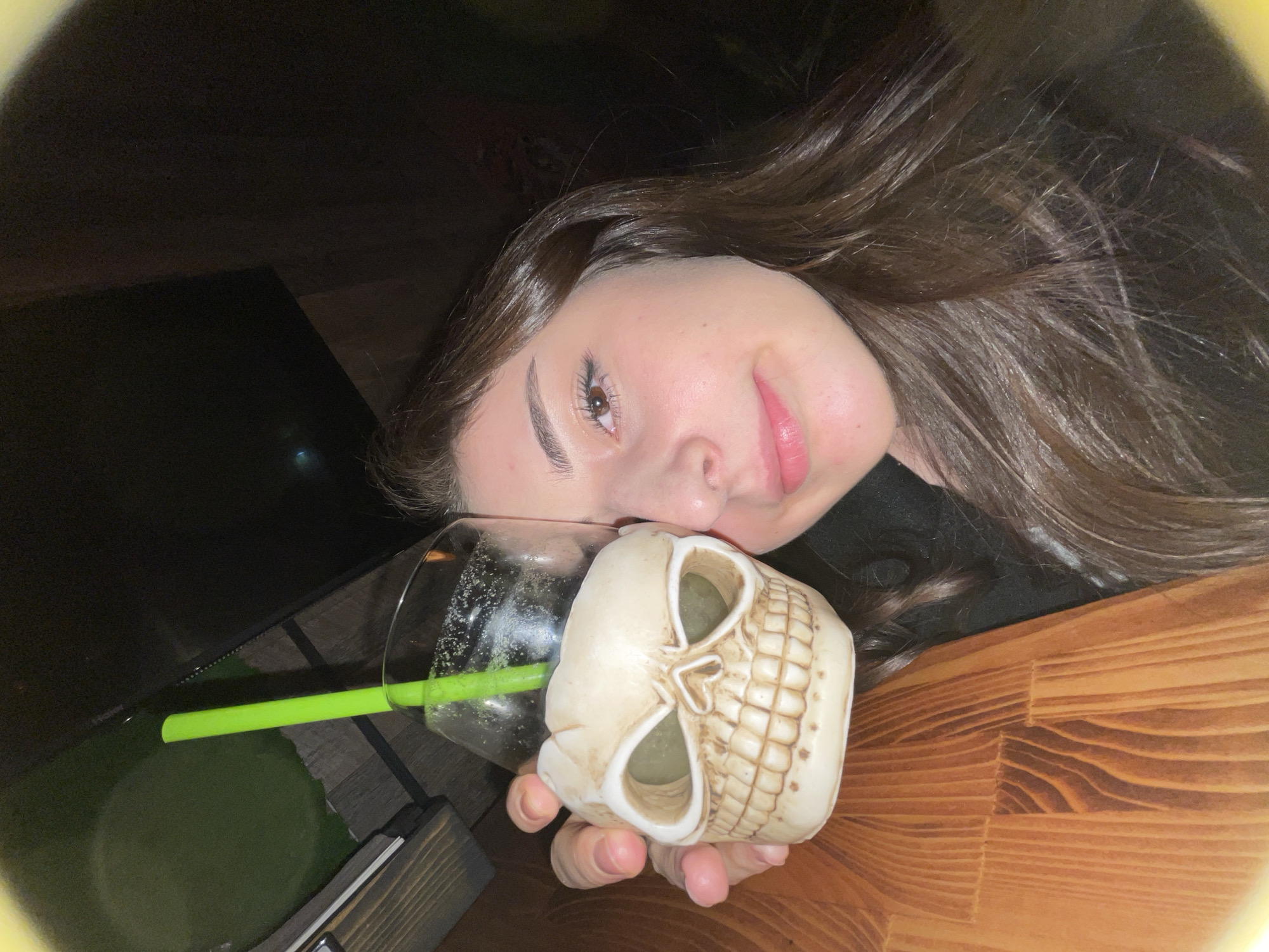 A woman with long brown hair is holding a glass in a skull-shaped holder with a green straw. The glass appears to contain a fizzy drink. The setting appears to be a bar or restaurant with a wooden table. The location cannot be determined to be Moscow, Russia without additional information.