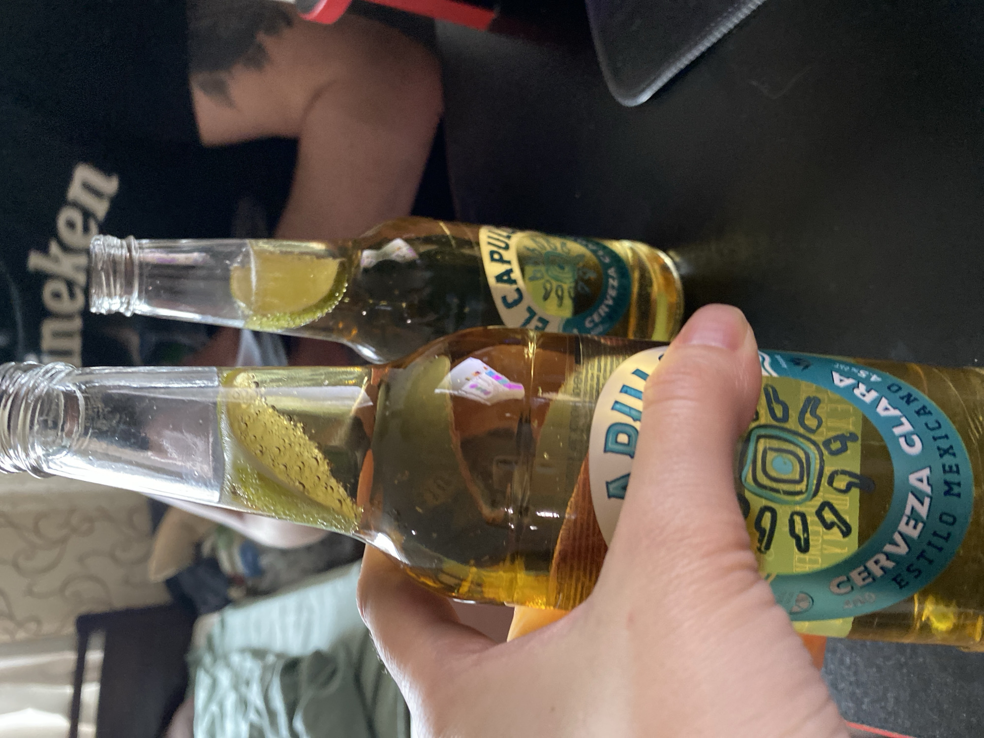 A close-up shot depicts two bottles of Mexican "Cerveza Clara" beer, each with a lime slice visible inside the neck. One bottle is being held by a hand. The background includes a black object possibly a monitor, a t-shirt with "Heineken" written on it, and a glimpse of a living room-like setting with a couch. The location appears to be indoors. No identifying features that would tie the photo to Izhevsk, Russia, are visible.