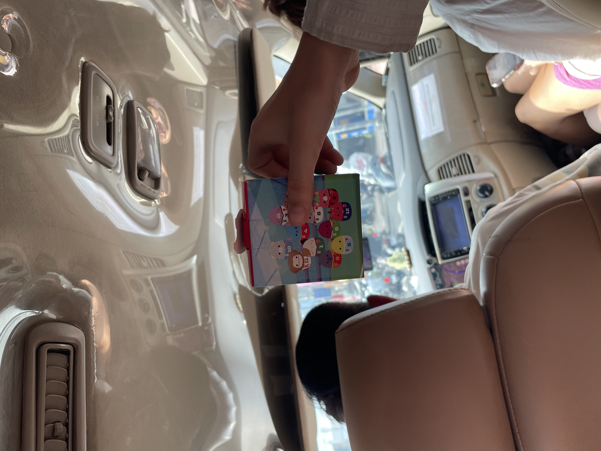 The image, taken in the city of Thủ Đức, Vietnam, depicts the interior of a vehicle, likely a car or van, with a focus on the ceiling and the upper portion of the dashboard and seats. The ceiling is light-colored and features air vents and overhead light fixtures. A hand is holding up a small, colorful box adorned with cartoon characters, possibly a collectible toy or a game. The box is positioned in the foreground, partially obscuring the view of the car's interior. Visible in the background are the heads of passengers in the front and back seats, their faces mostly obscured. The dashboard includes a navigation screen and various controls. Outside the vehicle, through the windows, a blurry urban scene is visible, with traffic and buildings suggested. The lighting suggests it is daytime, with natural light entering the vehicle.