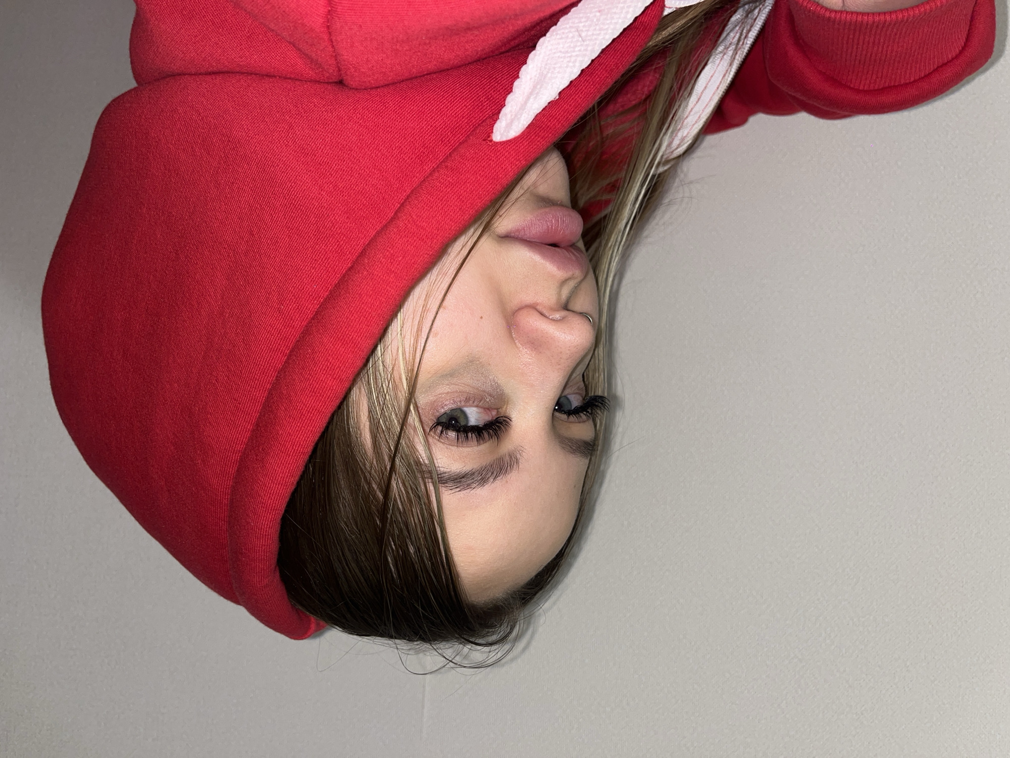 The media file presents a portrait of a young person wearing a bright red hooded sweatshirt. The image is oriented sideways, with the subject's head appearing on the left side of the frame, facing right, as if lying down or the camera was tilted.
The subject, appearing female, has fair skin and light brown hair with some lighter streaks, which partially covers their forehead and cheeks. They wear a red hoodie with the hood pulled up, partially obscuring the top of their head and ears. White drawstrings are visible around the hood's opening, contrasting with the vibrant red fabric. Their eyes, with notable long, dark eyelashes (likely enhanced), are open and look slightly upwards (relative to their head's orientation). Full, slightly parted lips and a small silver septum piercing on the nose are also visible. Their expression is neutral to slightly pensive.
The background is a plain, light gray wall, suggesting an indoor setting with even lighting. The red fabric of the hoodie shows a visible knit texture in the close-up crops. There are no other objects or discernible text in the image. The scene captures a close-up, informal moment. The location context provided is Krasnodar, Russia.