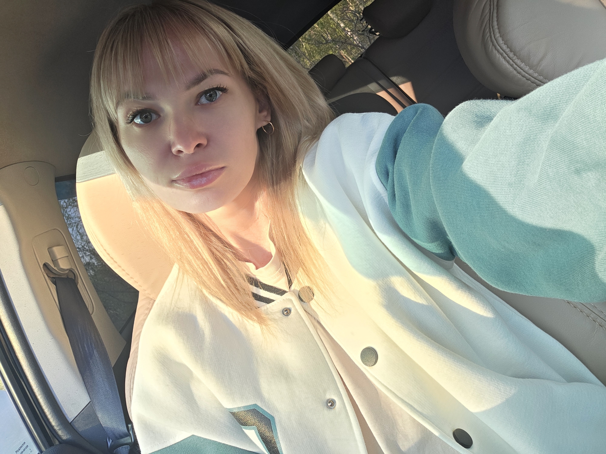 A blonde woman with fair skin sits in a car. She wears a white and blue bomber jacket, and small gold hoop earrings are visible. The car's interior features beige leather seats and a black seatbelt. Outside the car window, trees are visible, suggesting an outdoor setting. There is no information regarding the specific location of this image being taken in Chaikovsky, Russia.