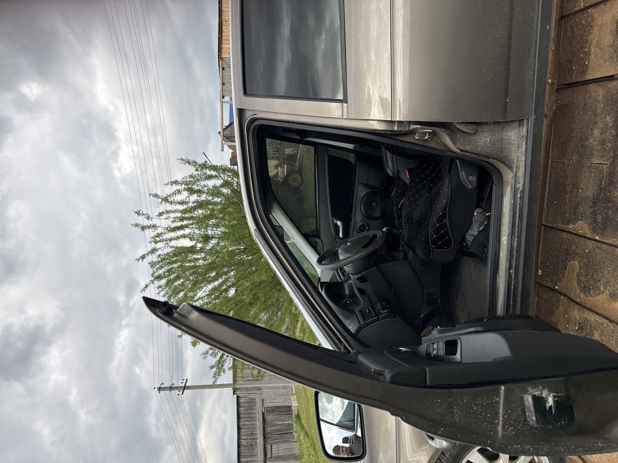 The vertically oriented image, appearing rotated 90 degrees clockwise, captures the side of a light-colored (possibly beige, gray, or light brown) SUV or crossover vehicle with its driver's door wide open. The scene is set outdoors, likely in a rural or semi-rural area of Бородулино, Russia.
Inside the car, visible through the open door, are parts of the dashboard, the steering wheel, and the driver's seat, which is covered with a black, quilted fabric. A speaker or vent is integrated into the door panel. The interior appears a bit dusty in places, particularly around the door frame and lower sections.
In the reflection of the car's side mirror, a man, presumably the photographer, is clearly visible. He has short hair and a beard or stubble and is holding a smartphone, indicating he is taking the picture.
The background on what would normally be the left side of an upright image shows an overcast sky, suggesting a cloudy day with soft, diffused light. Several power lines stretch across the sky, running horizontally. A tree with vibrant green, deciduous foliage is also prominent in this part of the scene. On the opposite side, what would be the ground or a structure, there's a weathered, rustic wooden fence or the facade of a wooden building, composed of planks or logs.
No specific activity is taking place; the car appears parked with the door ajar. No explicit text is visible in the image. The overall impression is calm, reflecting a typical day in the countryside under a cloudy sky.